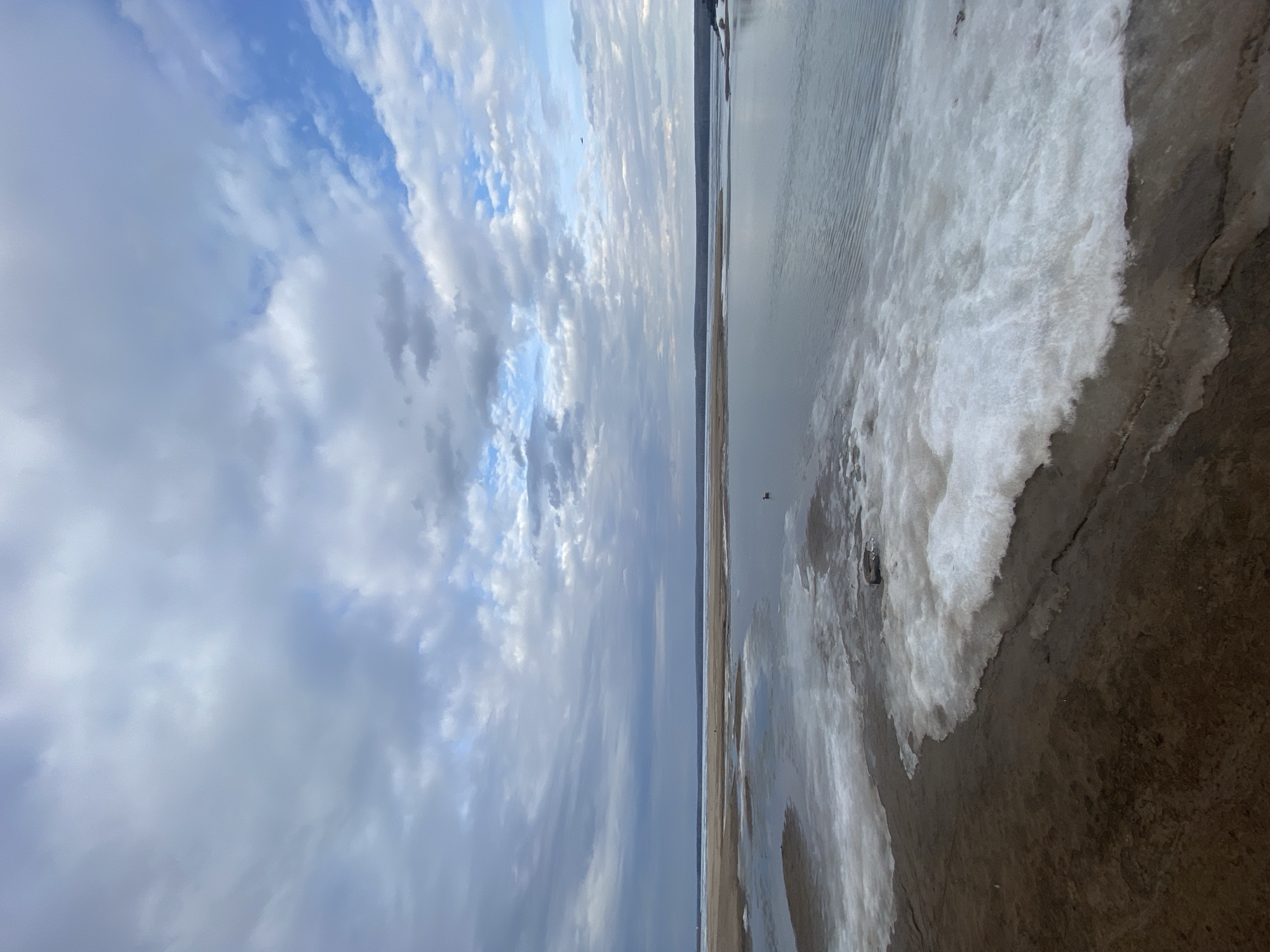 The image, viewed in its correct orientation (rotated 90 degrees counter-clockwise), presents a vast, open landscape composed primarily of sky, water, and shoreline.
**Subjects:** The upper half of the frame is dominated by a cloudy sky, featuring a mix of white and grey cumulus clouds against a pale blue background. The lower half depicts a wide body of water, possibly a lake or river, transitioning into a distinct shoreline. Along the immediate edge of the water, a significant amount of white, textured material, resembling ice or snow, is present, contrasting with the darker, muddy or wet sandy ground of the bank. A single, very small, dark, indistinct figure, possibly a person, is visible far in the distance near the horizon, seemingly standing on the water or ice. No other prominent objects are clearly identifiable.
**Scene and Setting:** The scene portrays a wide-open natural environment, characteristic of a large body of water with a spacious sky above. The setting, identified as Chaikovsky, Russia, is consistent with the presence of ice or snow along the shoreline, suggesting a cold climate.
**Activity or Event:** No specific activity or event is clearly depicted. The scene is static, with the distant figure being the only sign of human presence, contributing to a sense of calm and solitude.
**Notable Details:**
* **Time of Day:** The diffused lighting from the overcast sky suggests it is daytime, likely a cloudy morning or afternoon, without direct sunlight.
* **Weather:** The presence of clouds and ice/snow indicates cold weather conditions.
* **Location Cues:** The expansive water, distant horizon, and icy shoreline strongly suggest a large body of water, such as a major lake or river.
* **Emotions:** The overall impression is one of tranquility, vastness, and perhaps a touch of desolation.
* **Visible Text:** There is no visible text in the image.