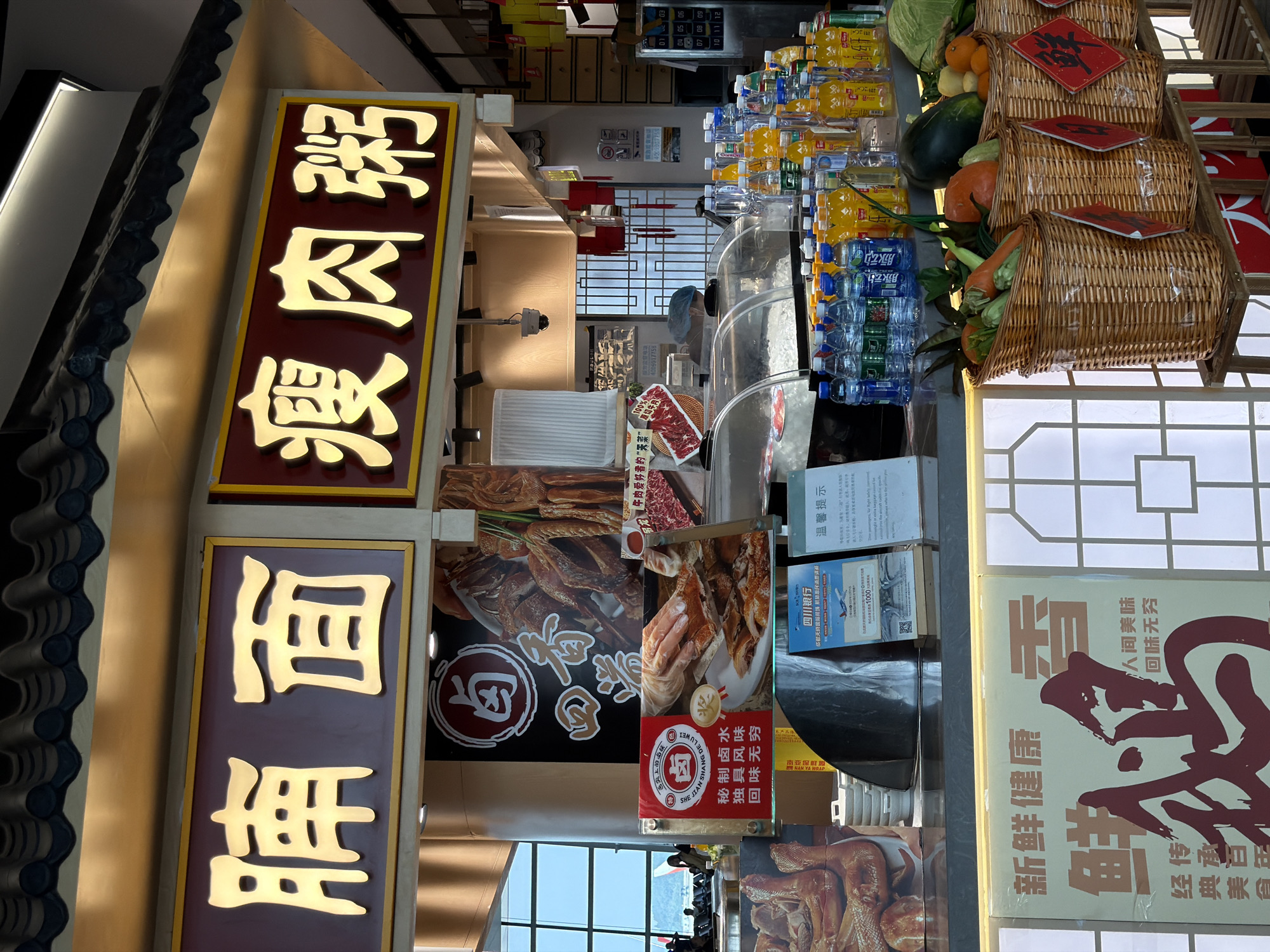 The image depicts a food stall, likely a restaurant or market stand, in Jianyang City, China. The stall sells braised meats and rice porridge, as indicated by the Chinese characters on the signs. The stall showcases a variety of products, including bottles of beverages and baskets filled with fruits and vegetables. The signage indicates promotions and offers, including a collaboration with the Sichuan Bank, offering 1000 yuan for consumers. The stall is likely located in a commercial area or market within Jianyang City.