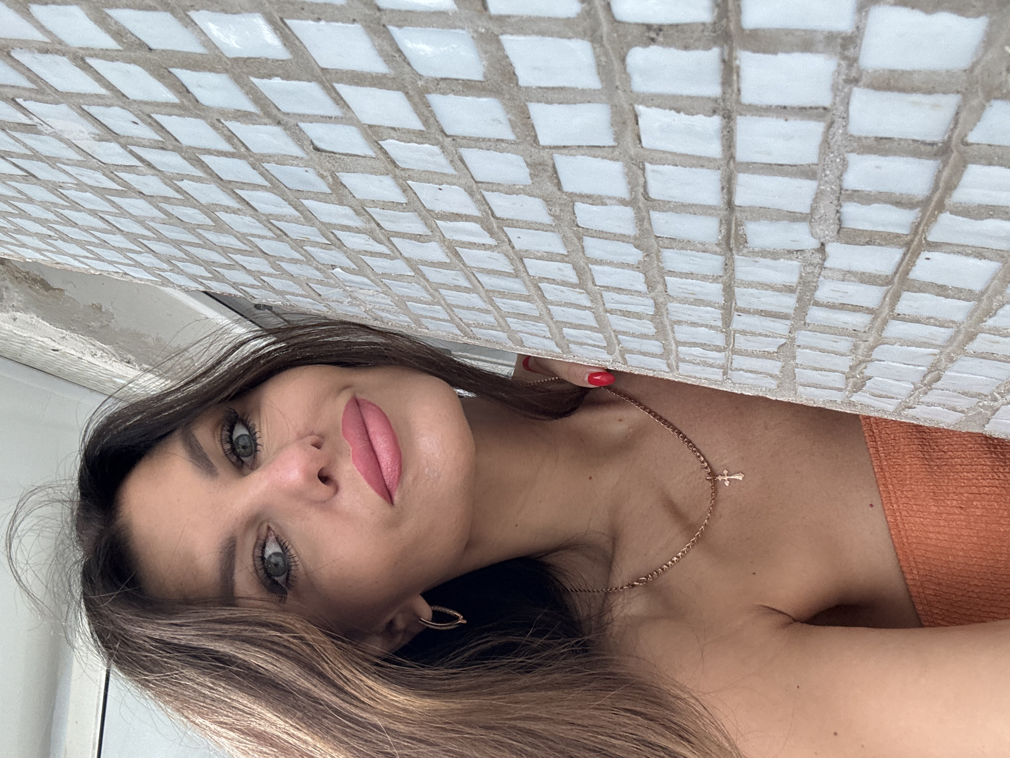 A medium close-up shot shows a woman with long, brown hair and blue eyes looking at the camera, resting her head against a tiled wall. She is wearing an orange top and a gold necklace with a cross pendant. Her lips are painted pink, and she has long eyelashes. Her right hand, with red nail polish, is touching her left cheek, near her ear. The tiled wall is white with grey grout and is positioned to the right of the woman, extending out of frame. The background is blurred and appears to be a window and ceiling. The image is undated and unlocated, and there is no specific information provided regarding the city of Chaikovsky, Russia.