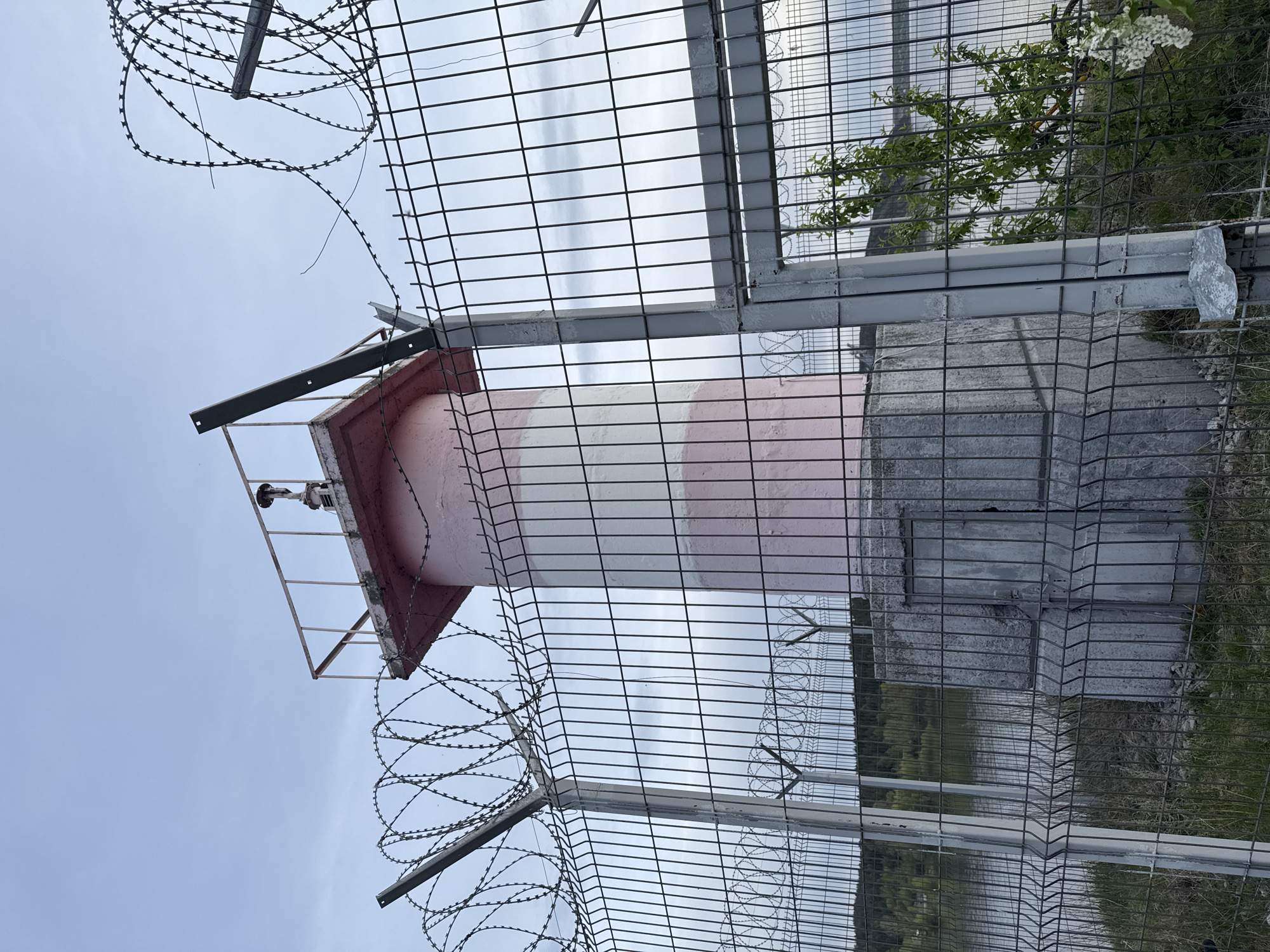 A low-angle, vertically-oriented outdoor shot depicts a small, striped lighthouse-like structure, enclosed by a fence topped with barbed wire. The structure features alternating pink and white horizontal stripes, and a dark red-roofed top section with a small observation platform. The fence is made of wire mesh. The background includes a body of water, trees and a cloudy sky. Based on the appearance of the structure and the information provided, it is likely that this image was taken in the city of Chaikovsky, Russia.