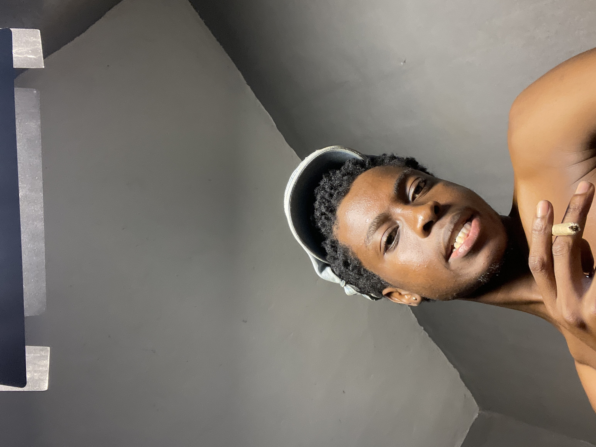 A portrait-oriented photograph captures a young individual with dark skin and short, dark curly hair, wearing a light-colored cap. The subject is looking directly at the camera with a confident and relaxed expression, featuring a slight smile that reveals their teeth. They appear to be shirtless, with a bare shoulder and upper chest visible on the right side of the frame.
The subject's right hand is raised near their face, and they are wearing a distinctive large, light-colored ring, possibly made of wood, on their ring finger. In their hand, they are holding what appears to be a hand-rolled cigarette or cigarillo, with a dark tip suggesting it might be lit or recently extinguished.
The background is minimalist, consisting of a plain, light grey wall or ceiling, indicating an indoor setting. Subtle architectural lines suggest a corner or junction in the room. A dark vertical element, possibly a door frame or furniture edge, is partially visible on the far left. The lighting is bright, illuminating the subject's face and creating highlights. The image is taken from a low angle and is significantly tilted. There is no visible text. The location context for this image is Nkpor, Nigeria.