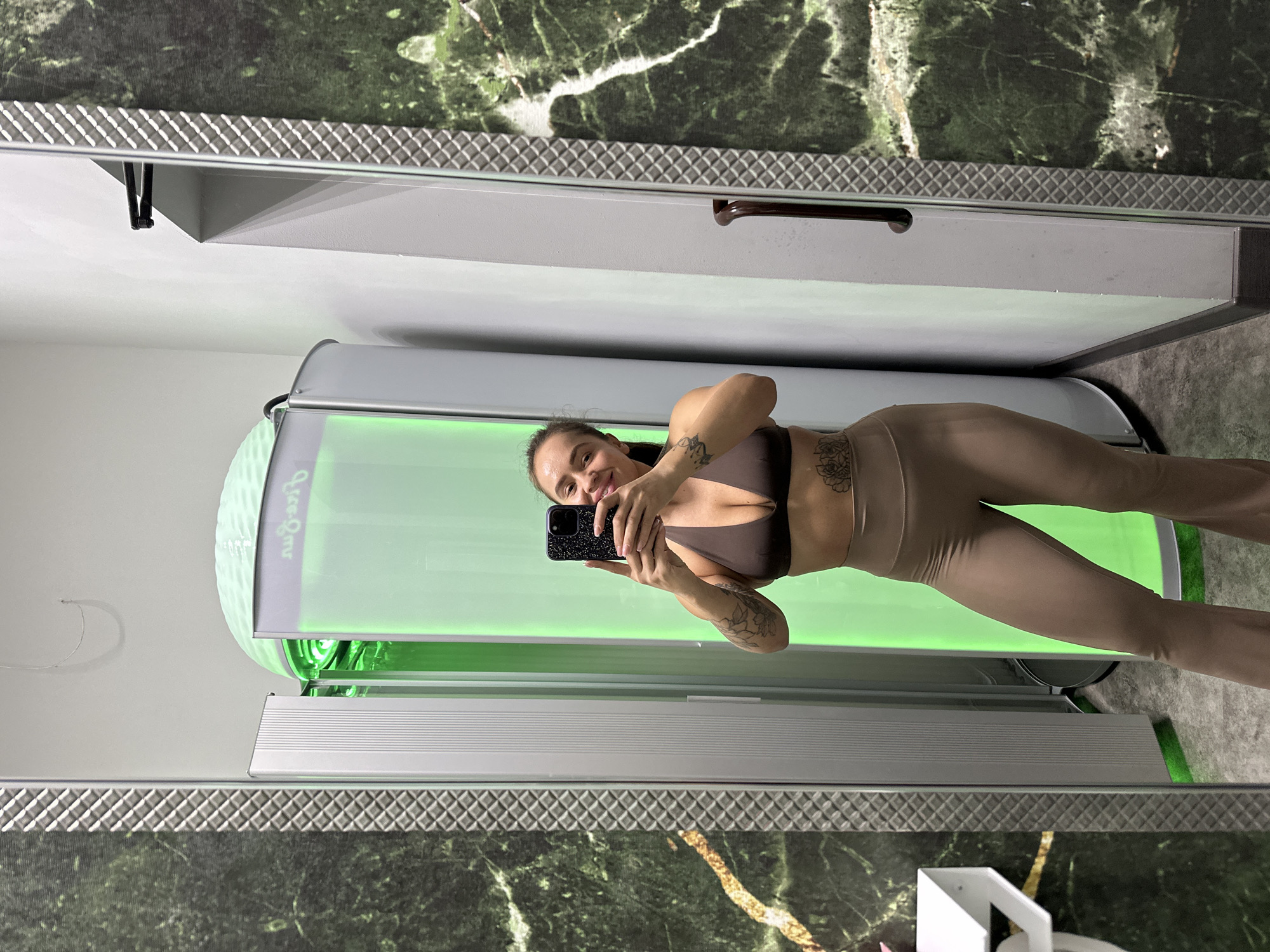 A woman with tattoos on her arms and abdomen is standing in a tanning booth and taking a selfie. She is wearing a brown bikini top and beige leggings. The tanning booth is illuminated with green light. The booth is located in a room with green marble walls and a mirrored wall. A white shelf is visible on the left side of the image.