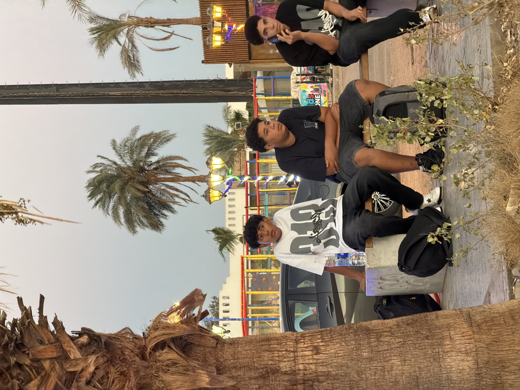 The image depicts three young males sitting on a bench outdoors, with palm trees and a building in the background. The setting appears to be a recreational area, possibly an amusement park or a promenade, given the colorful fencing and decorative lighting visible behind them. One of the boys is talking on a mobile phone, while the other two are looking away from the camera, seemingly engaged in their own thoughts. Bags are placed on the ground around them, and a bottle of water sits on the bench next to the boy on the left. The lighting suggests it is either early morning or late evening. The overall atmosphere is relaxed and casual. The location is specified as Sharm El Sheikh, Egypt.