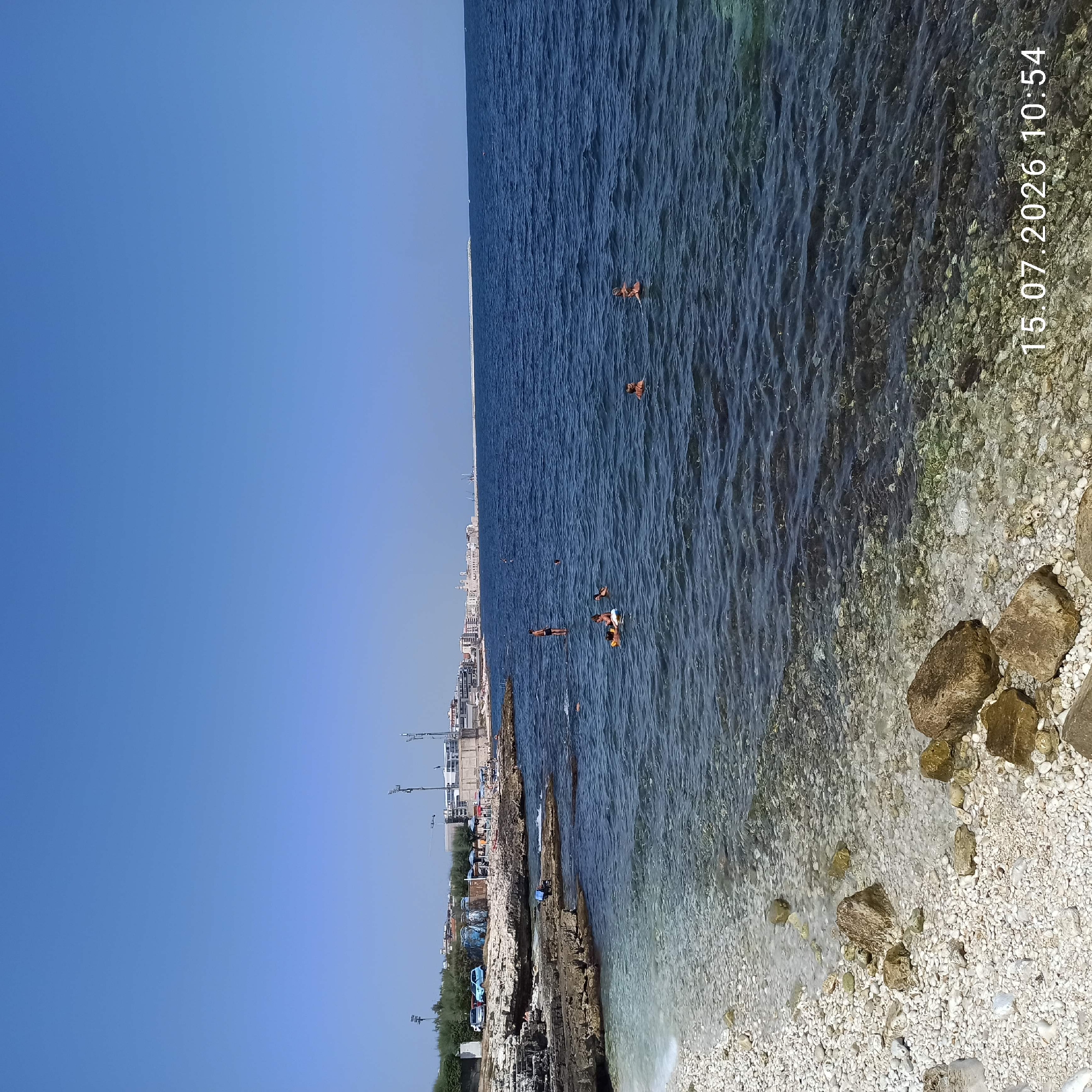 The image depicts a rocky coastline with clear, calm water and a bright blue sky. Several people are visible swimming and relaxing in the sea. In the background, a city skyline with buildings, trees, and light poles is present, suggesting an urban coastal setting. The date stamp on the bottom right indicates "15.07.2026 10:54". This scene is consistent with the coastal area of Molfetta, Italy, known for its rocky shores and proximity to the city.