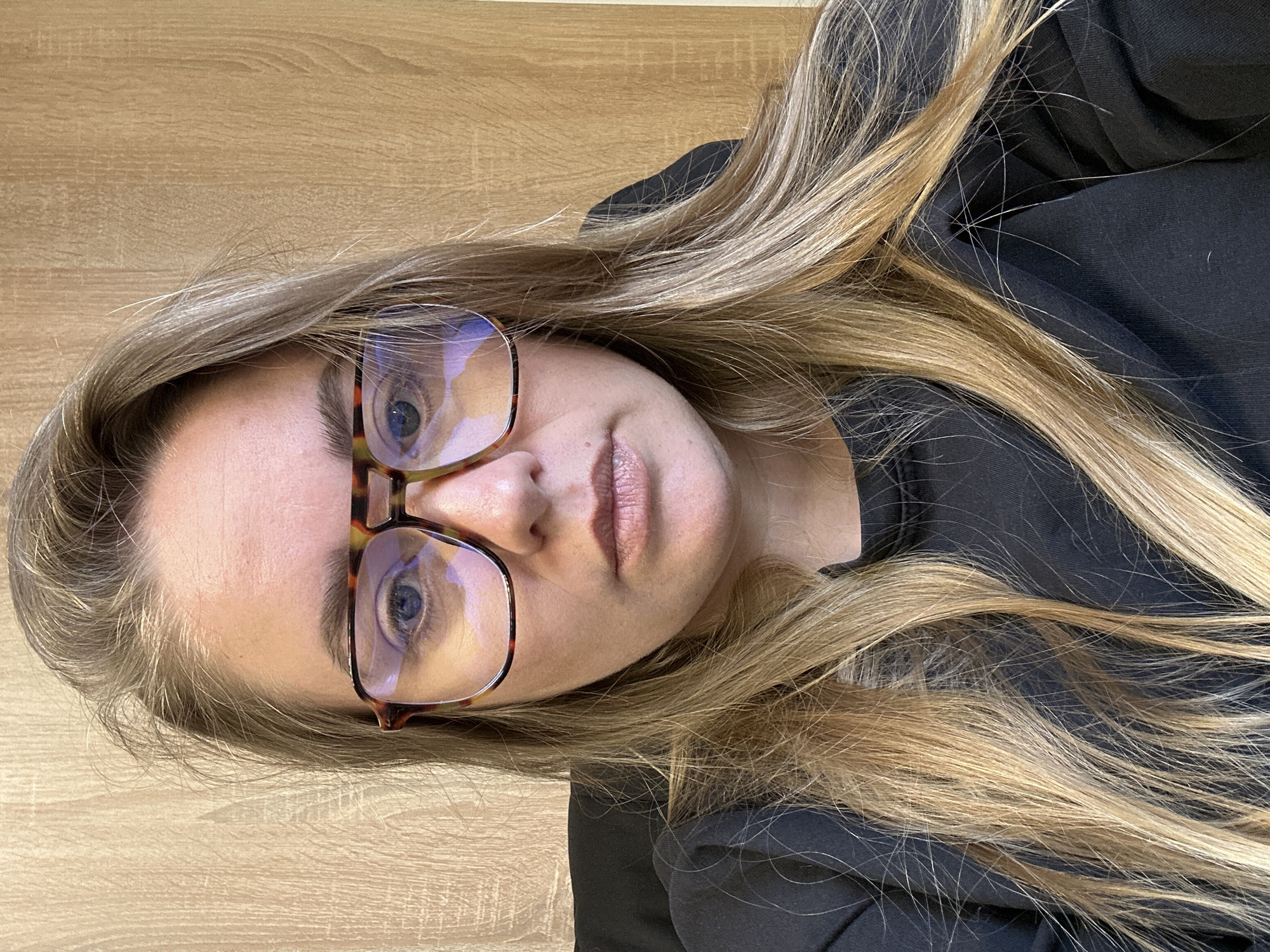 A woman with long blonde hair and blue eyes is wearing tortoise-shell patterned glasses. She is wearing a black top. The background appears to be a light-colored wooden surface. There is no indication of Ural'skoe, Russia in the image.