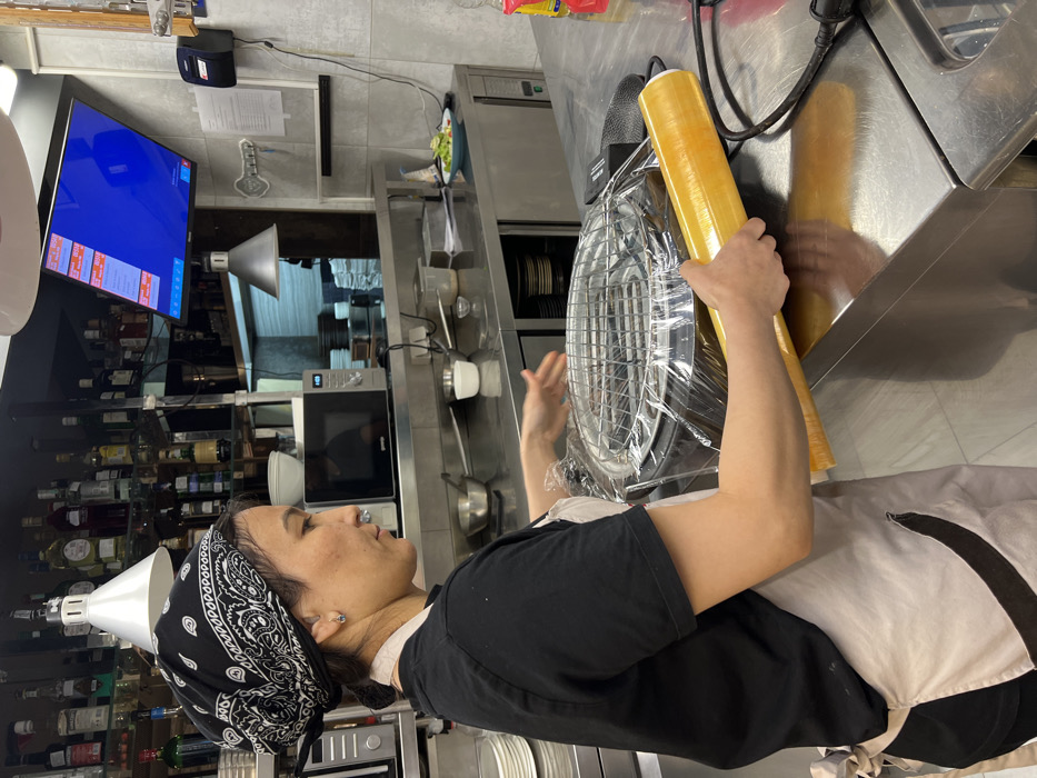 A person in a restaurant kitchen wraps an electric heater in plastic wrap. The individual wears a black t-shirt, apron, and a bandana on their head. Shelves filled with bottles are in the background, along with stainless steel equipment. A monitor displays a menu or order list on the wall.