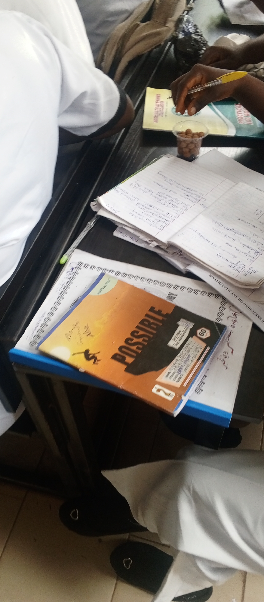 This image captures a close-up, angled view of a group of people engaged in what appears to be a study session at a school or educational institution. The primary subjects are hands and arms, with one person actively writing with a yellow pen in a notebook filled with handwritten text. Another notebook with the word "POSSIBLE" prominently displayed on its orange cover is also visible, alongside other papers and books. The setting is a dark table surface, suggesting an indoor environment.
The activity is clearly focused on learning or taking notes, as indicated by the writing and the presence of educational materials. The lighting suggests it could be daytime, but there are no specific cues to determine the exact time. The overall mood appears to be one of concentration and academic pursuit.
Notable details include a small plastic cup filled with what look like small, round snacks. Text visible on the notebook covers and pages includes "POSSIBLE," "PRACTICAL LOGBOOK," "60," and some handwritten notes in what appears to be English. There are also some symbols and what might be a school logo on one of the notebooks. The image also shows parts of white clothing, possibly uniforms, and dark footwear, hinting at the attire of the students. The perspective is from above and slightly to the side, looking down at the table.