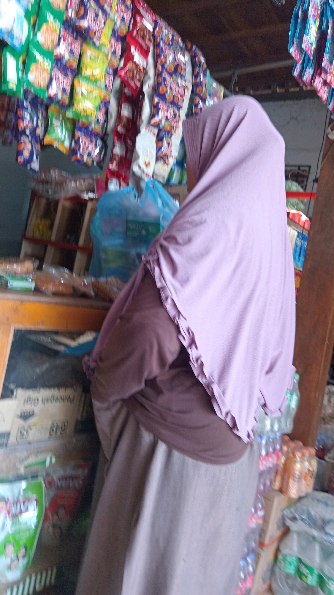 In Kedungwaru, Indonesia, a woman wearing a light purple hijab and a brown top stands in front of a stall displaying various snacks. The stall is adorned with colorful packets of snacks hanging from above. To the right of the woman, a stack of plastic bottles, some filled with beverages, is visible. The background shows shelves stocked with more items, suggesting a small shop or market environment.