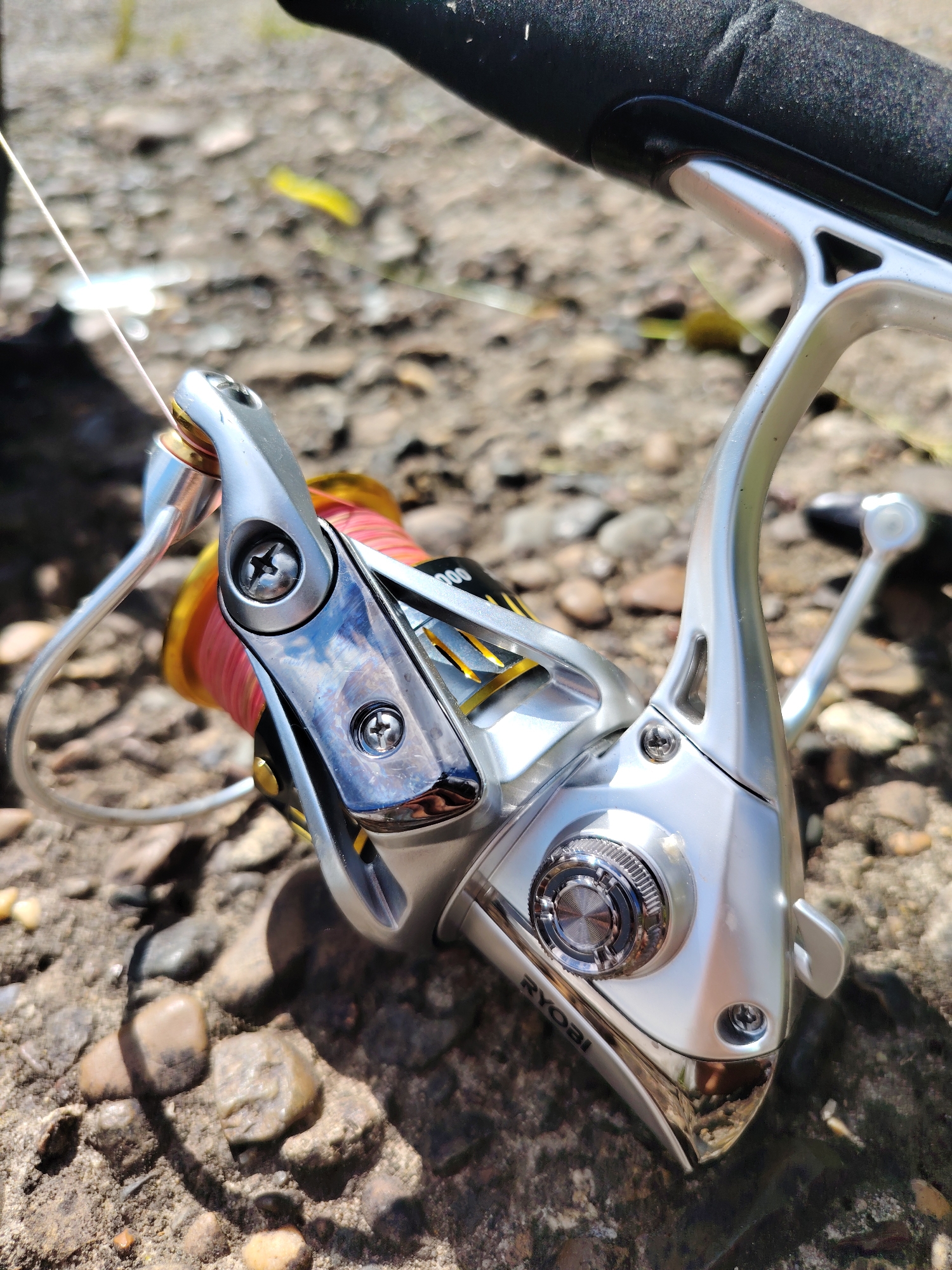 The image depicts a close-up of a Ryobi fishing reel mounted on a fishing rod, resting on a surface covered with small stones and debris. The reel is silver, with visible screws, a spool with red fishing line, and a handle. The fishing line extends from the reel's bail. The rod appears to be black. The background suggests an outdoor setting with natural light. The location in the city of Chaikovsky, Russia, cannot be determined from the image.