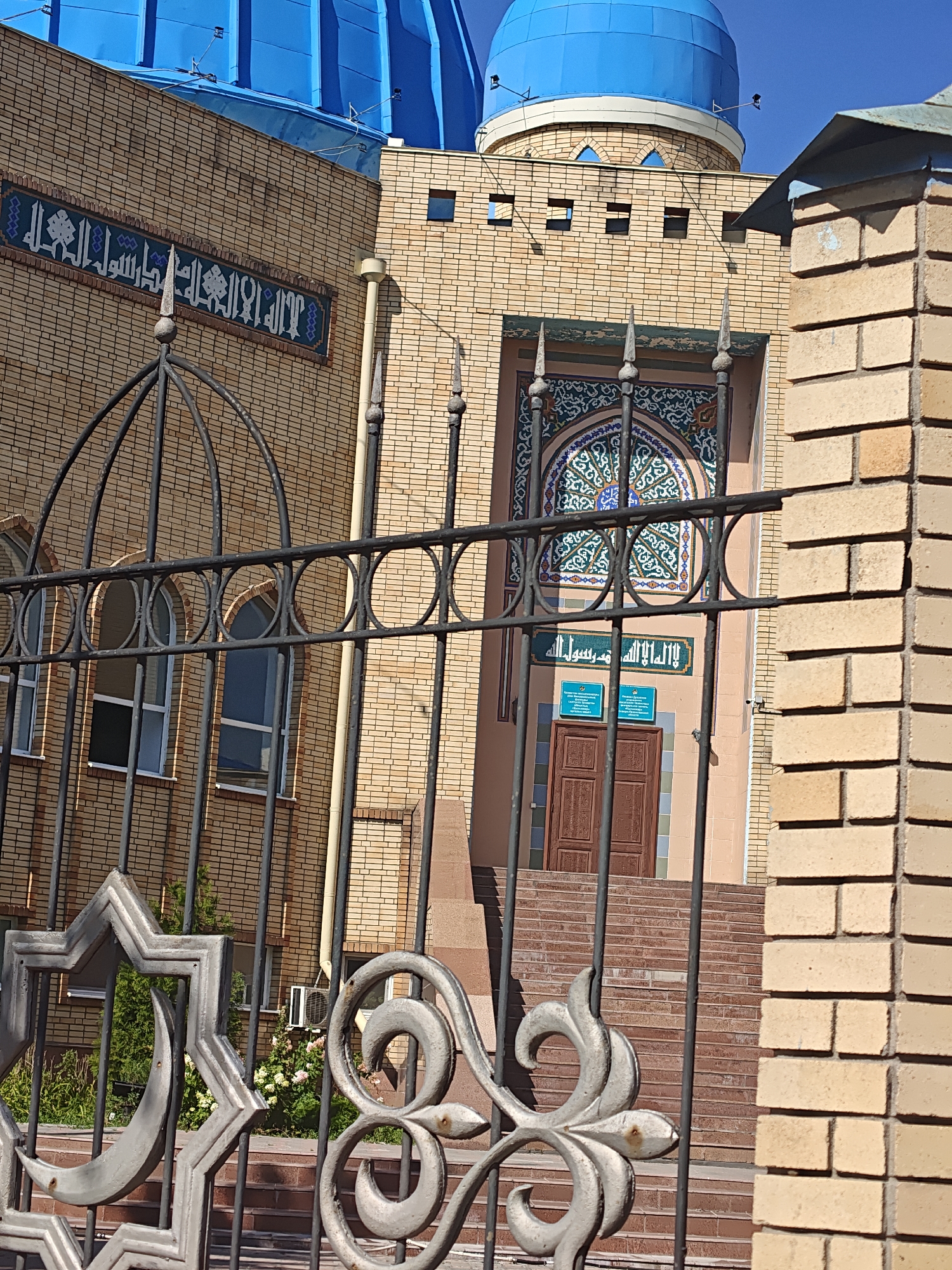The image shows the exterior of a mosque in Petropavl, Kazakhstan. The mosque features light brown brickwork and two prominent blue domes. Decorative Islamic calligraphy is visible on the facade, with a colorful mosaic arch above an entrance. A decorative black metal fence with star and crescent motifs partially obscures the view of the building's lower levels, including arched windows and a wooden door. Steps lead up to the entrance. The scene is illuminated by sunlight, casting shadows on the building and fence. No people are visible in the frame.