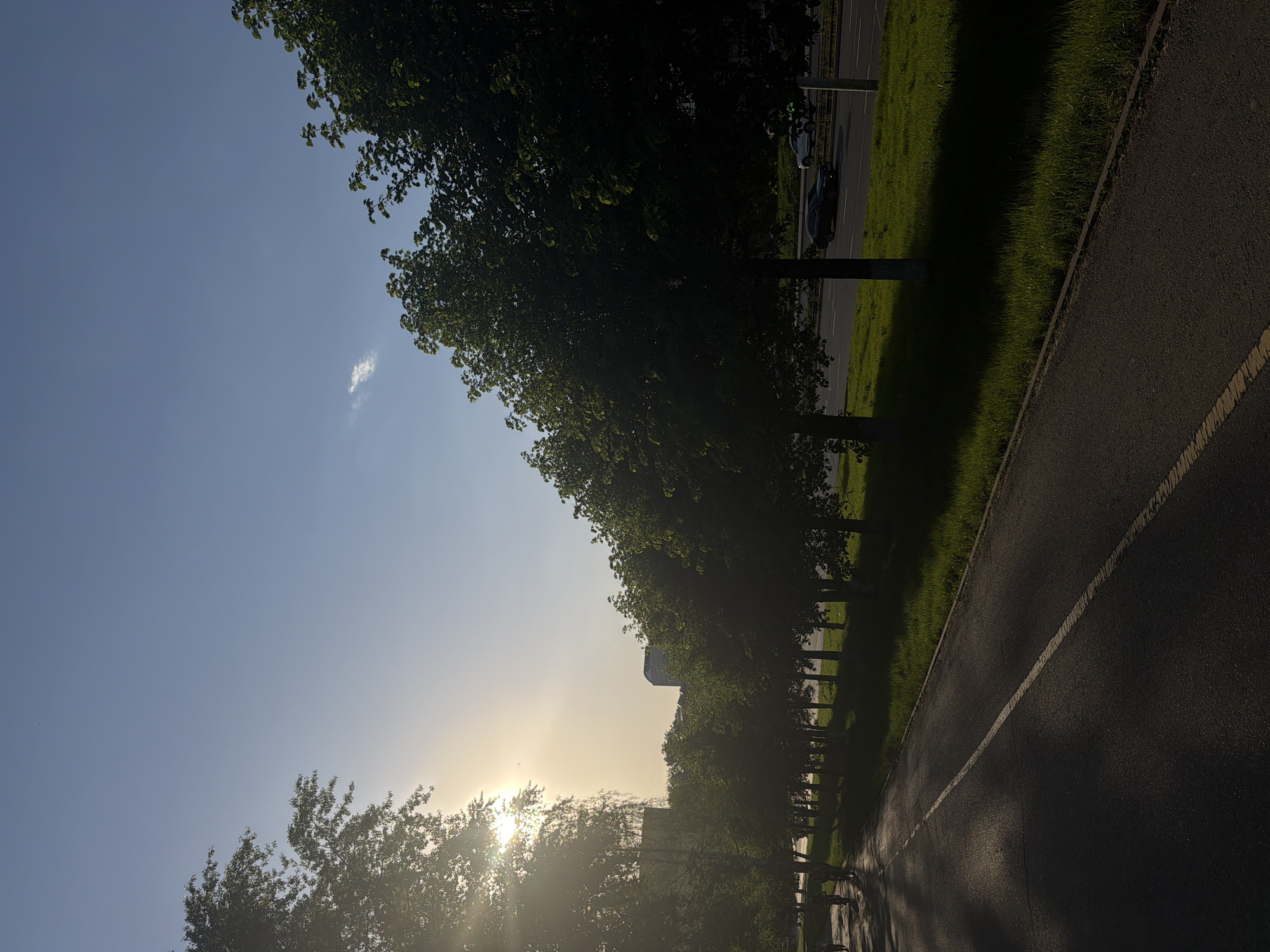 The image depicts a scene in Izhevsk, Russia, taken during sunrise or sunset. A paved road with a white line marking separates it from a strip of green grass. Trees line the left side of the road. In the distance, buildings are visible, obscured by the sun's glare. Sunlight streams through the trees. The sky is a clear blue transitioning to a lighter, sun-drenched horizon. There are no people visible in the image.