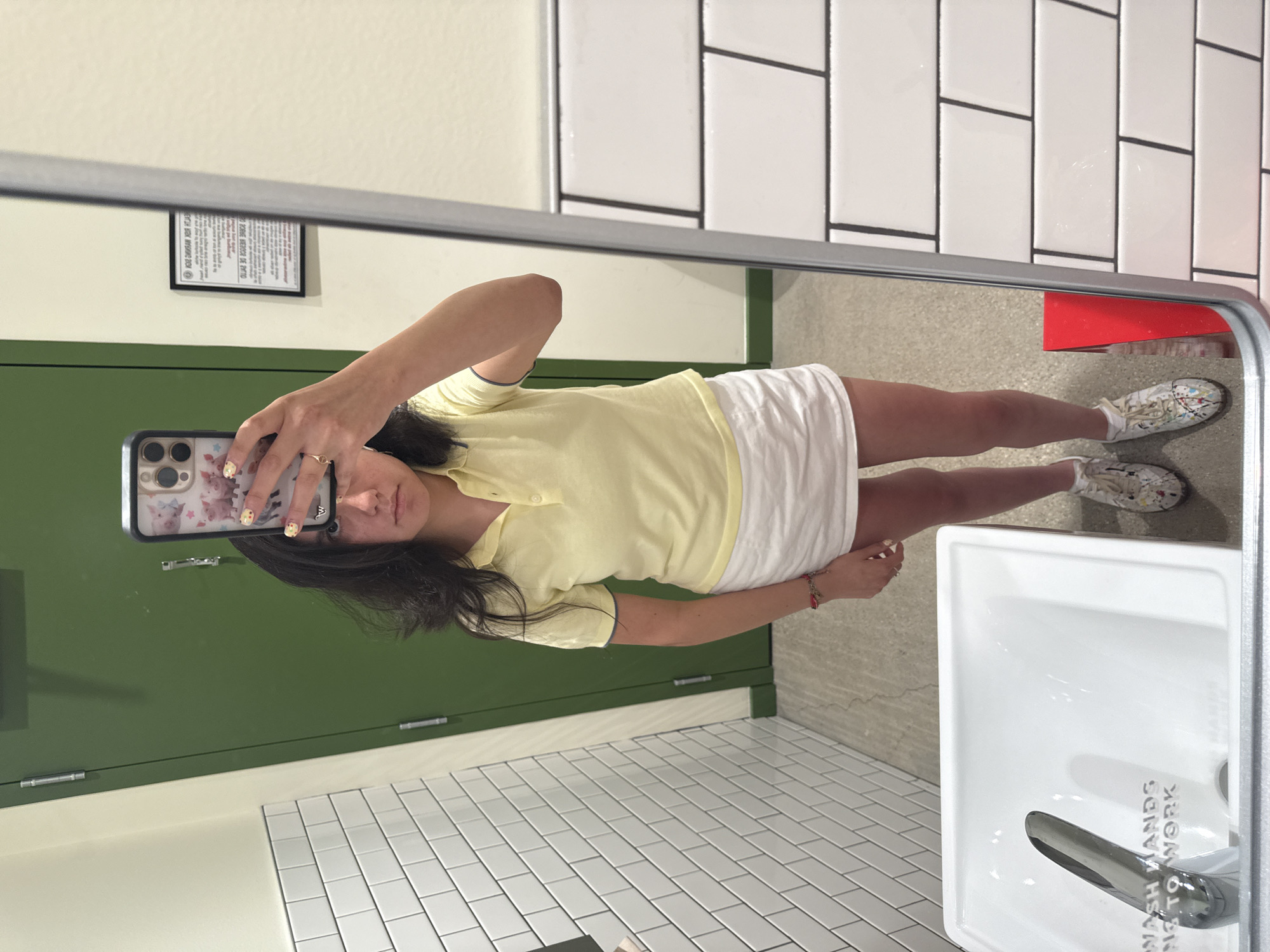 A mirror selfie reveals a woman in a public restroom. She is wearing a yellow polo shirt and a white skirt, with patterned sneakers on her feet. The background features a green door, white tiled walls, a stainless steel framed mirror, and a white porcelain sink. The sink has an inscription that says "WASH HANDS, BACK TO WORK." Based on the style and design of the restroom, it is likely located in Austin, Texas.