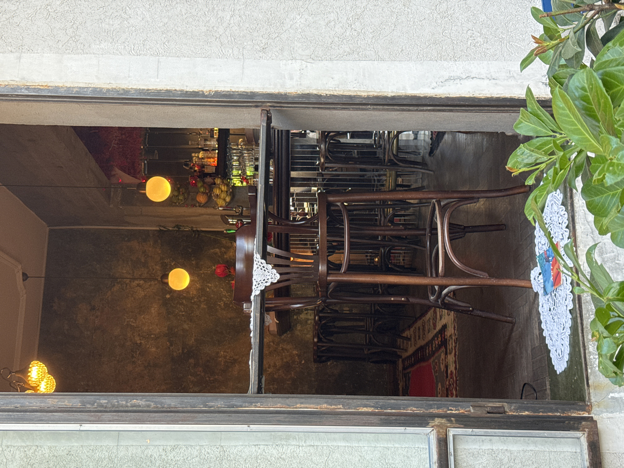 The image, taken at eye level, depicts a view through a window into what appears to be the interior of a bar or restaurant. The location is likely Belgrade, Serbia.
Inside, several dark wooden chairs are stacked on top of tables, suggesting the establishment is either closed or undergoing preparations. Soft, warm lighting is provided by multiple pendant lamps, casting a glow on the dark-toned walls. A display of bar items is visible in the background.
The window frame itself is aged and somewhat worn, and there are plants visible growing on the outside of the building. A crocheted doily is placed on the window ledge.
