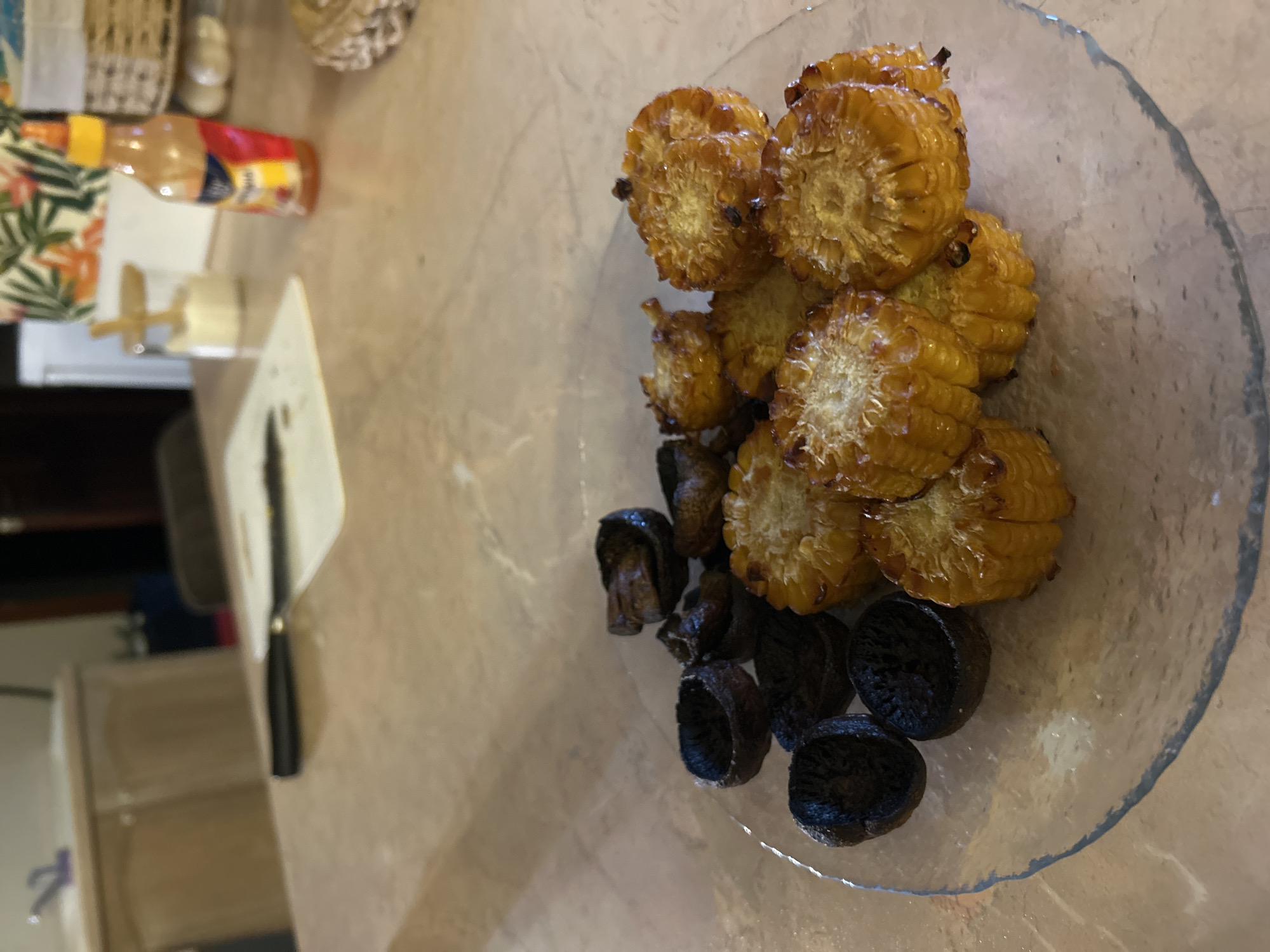 The image depicts a clear glass plate containing grilled corn on the cob, cut into segments, and what appear to be grilled mushrooms. The plate is placed on a light brown, possibly marble or granite, surface. To the left of the plate, there is a serving utensil (spatula) and additional objects including a bottle of hot sauce and other kitchen items. The location information, Chaikovsky, Russia, is not discernible from the visual information provided. The scene is indoors.