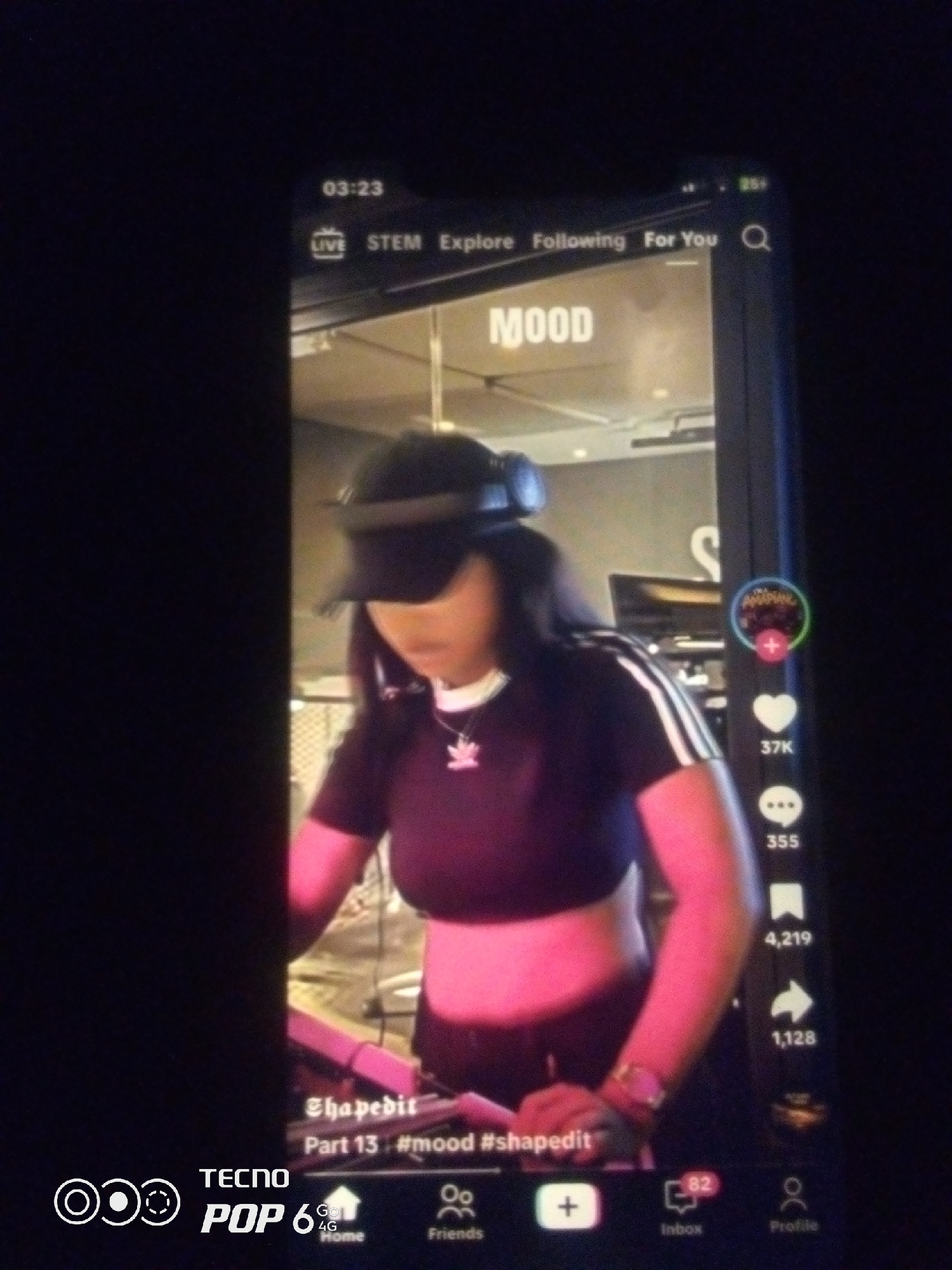 A vertical, high-angle shot captures a dimly lit smartphone screen displaying a social media feed. The feed features a woman wearing a black baseball cap, headphones, a black crop top with white stripes on the sleeves, and a silver watch on her left wrist. She is looking down and appears to be operating DJ equipment. The screen shows video playback controls, a "MOOD" text overlay, a username "ShapedIt", and hashtags "#mood #shapedIt". Social media engagement metrics such as likes (37K), comments (355), and shares (1,128) are visible on the right side of the screen. The bottom navigation bar displays icons for Home, Friends, Inbox (with a notification count of 82), and Profile. The phone itself appears to be a Tecno Pop 6. The lighting is predominantly from the phone screen, creating a dark environment around it. There is no discernible city or specific location within Howayi, Nigeria, or any other city depicted.
