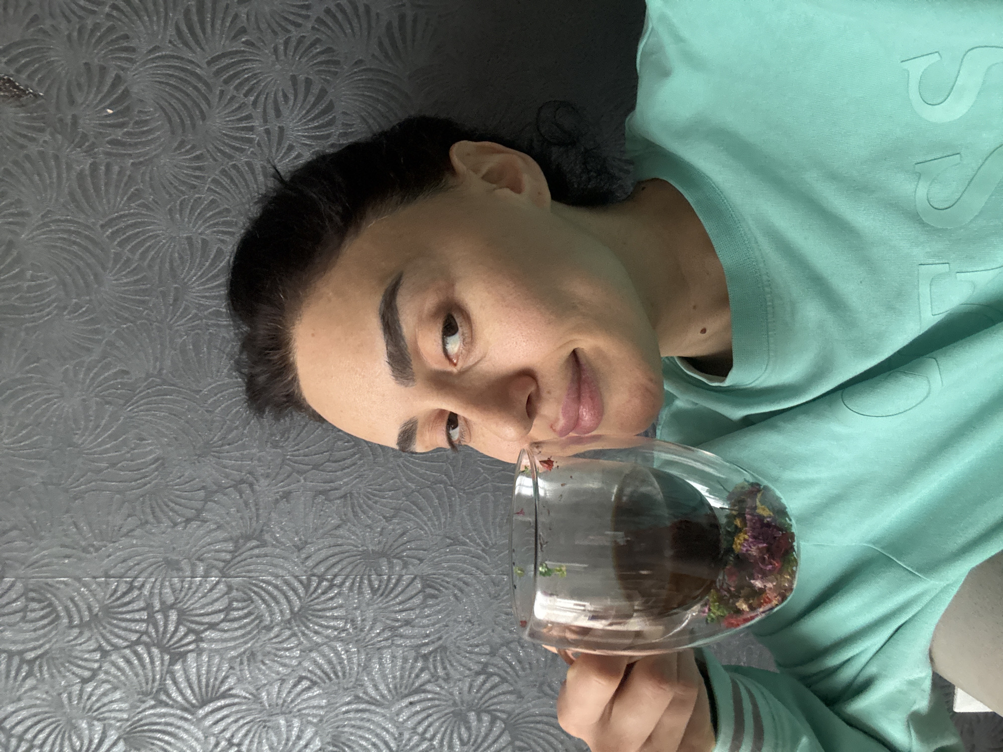 A person is lying on their left side on a gray patterned surface, wearing a light green sweater. They are holding a clear glass containing a dark liquid and colorful plant matter. The person has dark hair and brown eyes. The location is not discernible from the image.