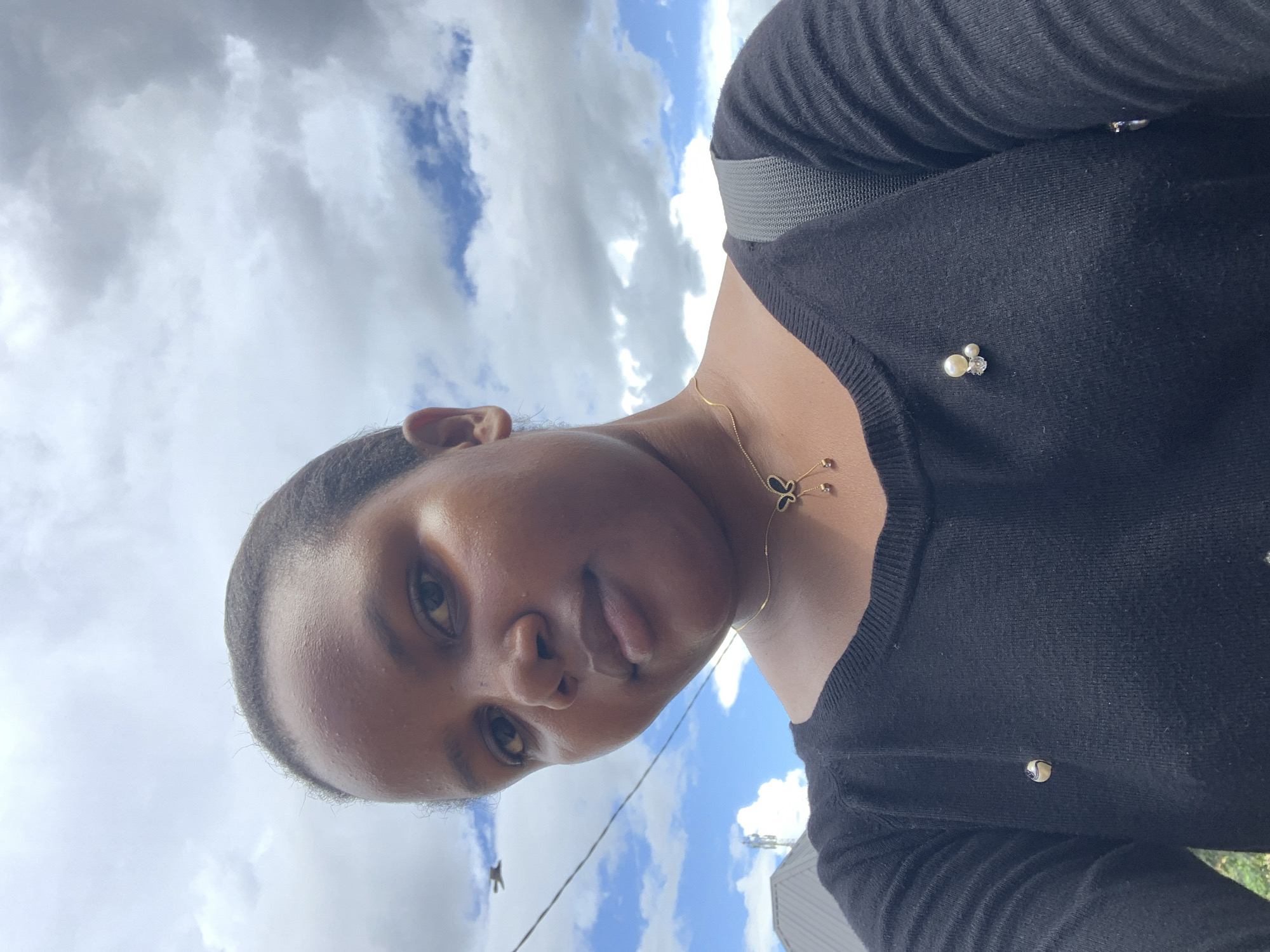 An outdoor, upward-angled portrait depicts a female individual with dark brown skin and short, dark hair, looking directly at the camera with a neutral expression. She wears a black long-sleeved knitted top. A gold-colored necklace with a black and gold butterfly pendant from which three dark beads are suspended is visible on her chest. A brooch consisting of two white pearls and a clear gemstone is pinned to her upper right chest (viewer's left), and a small round white pin is on her lower left chest (viewer's right). A grey backpack strap is visible over her right shoulder (viewer's left). The background features a partially cloudy sky with sections of blue visible through white and grey clouds. Distal elements include a partial view of a grey sloped roof and a communication tower structure on the left side. A thin black wire crosses the upper left, and a bird in flight is visible below it. The scene is located in Dar es-Salaam, Tanzania.