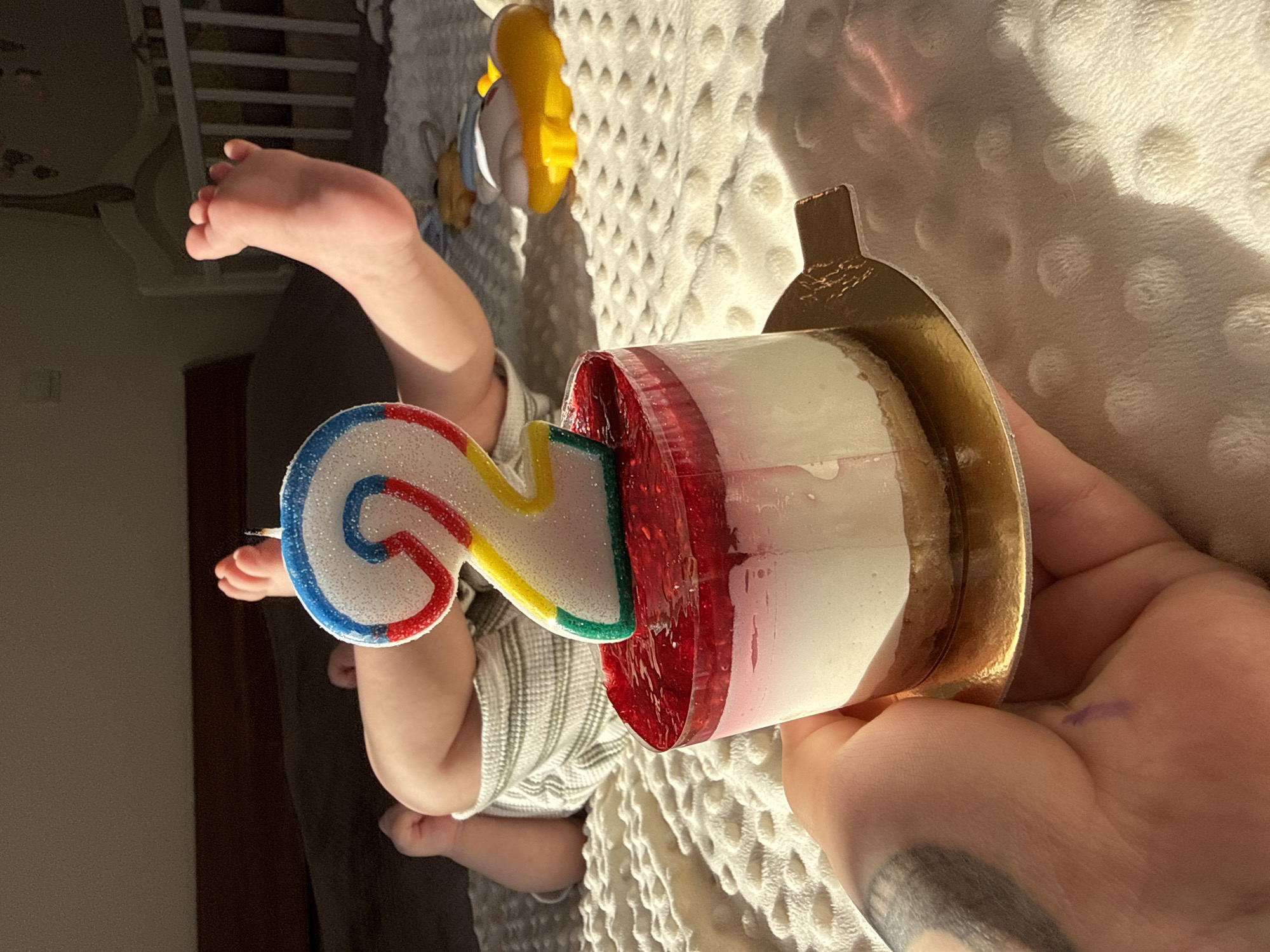 A partially-visible baby lies on its back on a textured white blanket, with a foot extending upwards. A small, decorative cake with a "2" candle is held in the foreground by a hand with a tattoo. A stuffed animal is visible in the background. The location appears to be indoors, possibly a home. There is no evidence to suggest the location is in Bar, Montenegro.