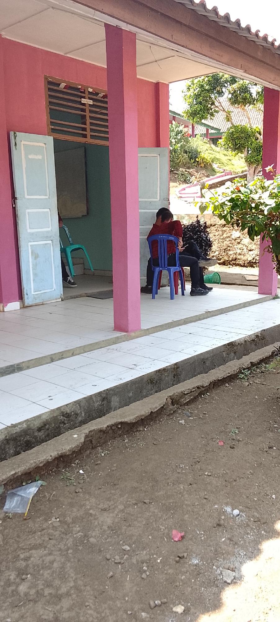The image depicts an outdoor scene at a building with pink walls and a white tiled walkway. Two individuals are present. One person, wearing a red and black checkered shirt and black pants, is seated on a blue plastic chair. The other person, wearing a red shirt and black pants, is seated on a red chair and appears to be interacting with the first person. A green plastic chair is visible inside a doorway. In the foreground, there is a concrete drainage ditch with some debris, including a clear plastic bag with a yellow handle. Beyond the building, there are trees and vegetation, with a sloped area in the background. The overall environment suggests a somewhat informal setting, possibly in a rural or semi-urban area, given the presence of outdoor seating and natural elements. The location is specified as Tanjung Setia, Indonesia.