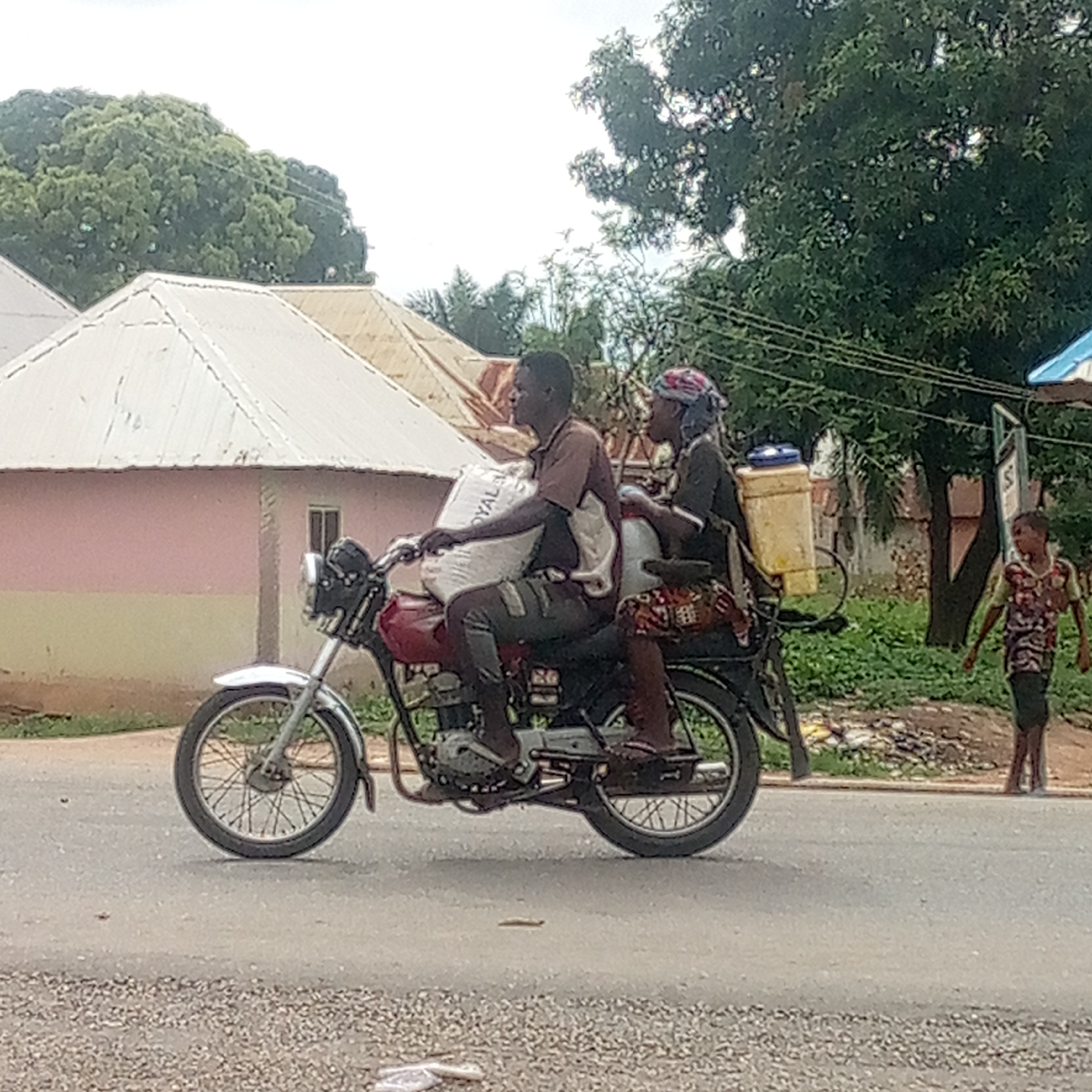 In Mararaba, Nigeria, a man and a woman are riding a red motorcycle down a paved road. The man drives, holding a large white sack in front of him. The woman sits behind him. A yellow container and other items are on the motorcycle. A child walks on the side of the road. Buildings and trees are in the background.