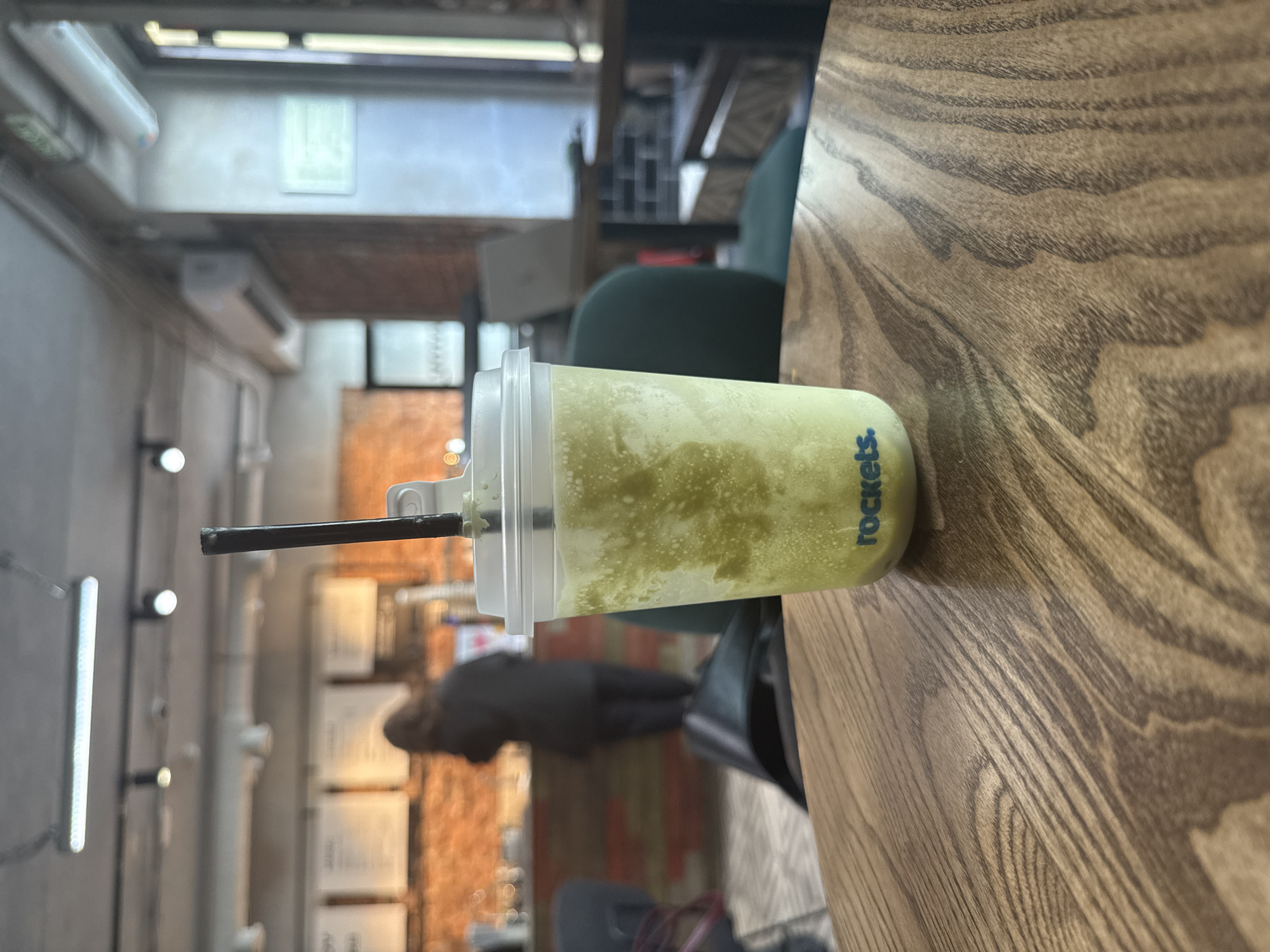 A partially filled, clear plastic cup with a green smoothie sits on a wooden table. The cup has a lid and a black straw. The label on the cup reads "rockets." The table has a distinct wood grain pattern. In the background, there is a blurred interior of a cafe or restaurant with neutral colors and brick walls. People are moving around in the background. The precise location in Moscow, Russia, cannot be determined from this image.