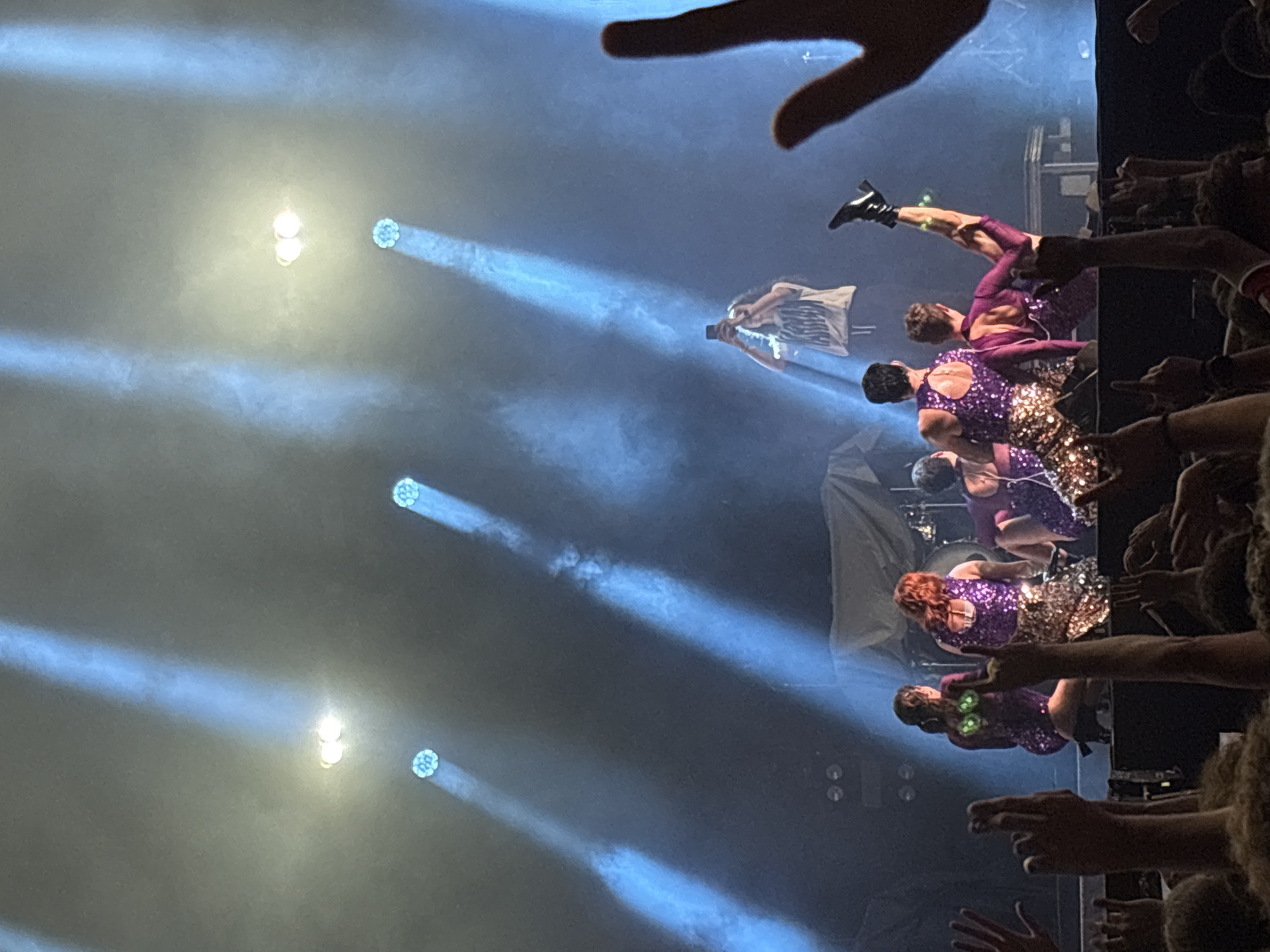 A low-angle, medium-shot image shows a band performing on stage, captured from the perspective of an audience member. The performance features four dancers or singers wearing glittering costumes in shades of purple and gold, performing choreographed movements. Light effects are visible in the background, with multiple spotlight beams illuminating the stage area. Audience members' hands and arms are raised towards the stage, capturing photos or videos. The location is not evident in the image.