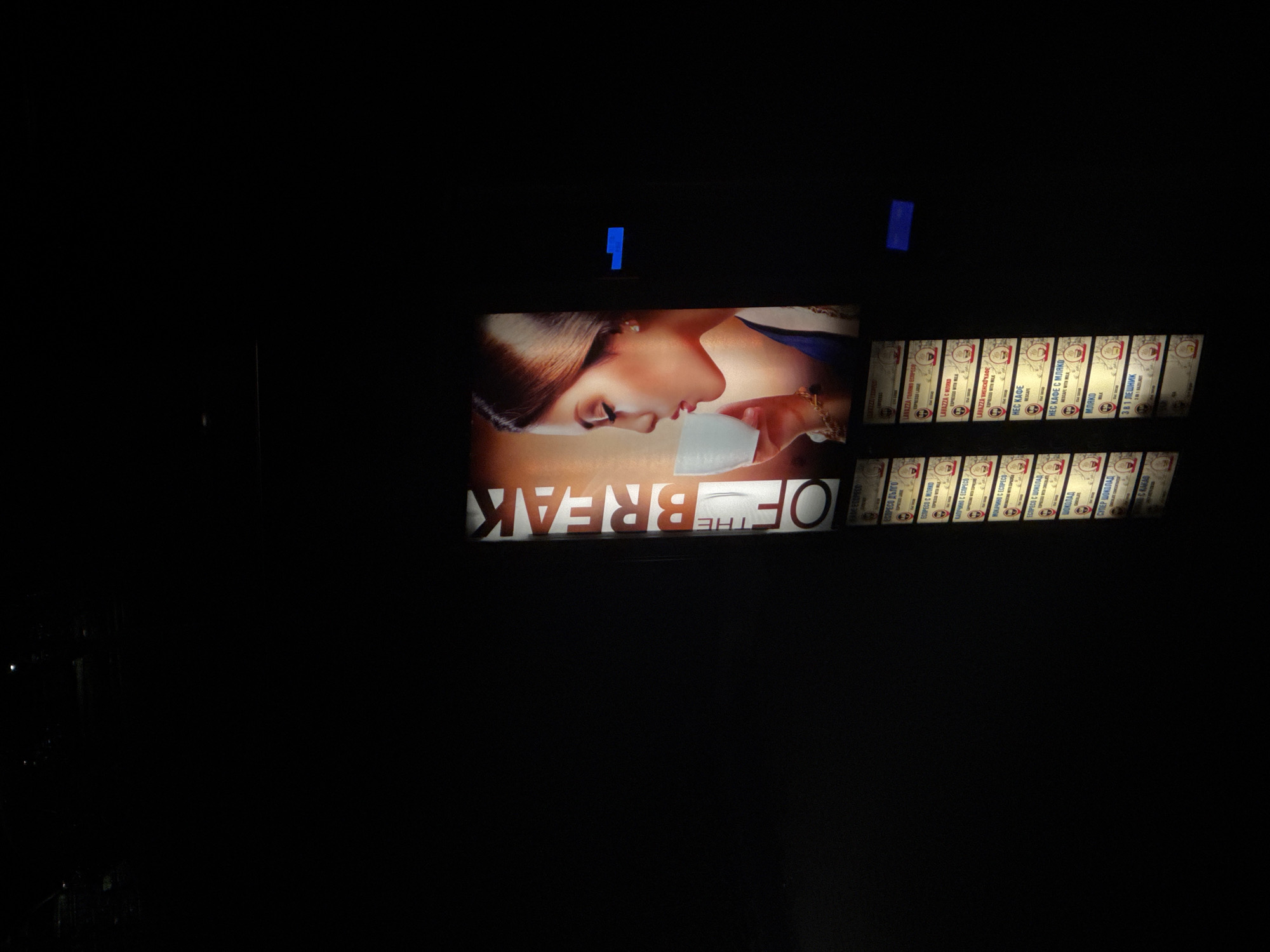 The image depicts a dimly lit interior, likely a kiosk or small establishment in Varna, Bulgaria, at night. On the right side is a menu board displaying various coffee options, with the names of the beverages written in Bulgarian. Some options include "Еспресо Дълго," "Еспресо с мляко," and "Нес Кафе." The text "Lavazza" is visible on some of the menu items. To the left of the menu board, an illuminated advertisement displays the words "THE OF BREAK" and a woman drinking coffee. Two small blue strips are placed above the advertisement. The surroundings are dark, with minimal visible details beyond the illuminated elements. The scene appears to be shot from a low angle, emphasizing the verticality of the display.