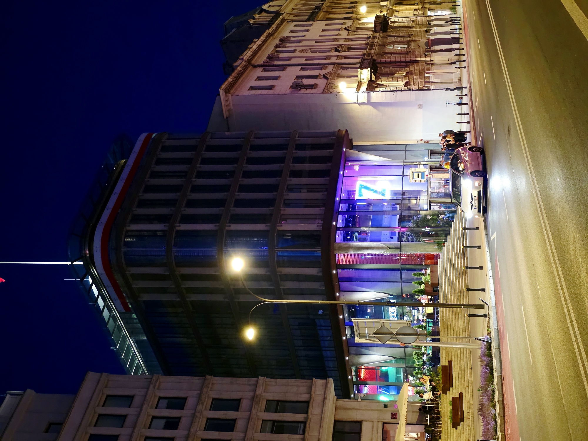 An outdoor, low-angle, nighttime, long shot captures a street scene in Warsaw, Poland, dominated by the modern, glass-fronted facade of the "7" building and an adjacent older building. The "7" building, illuminated with vibrant blue and purple lights, features a prominent, illuminated "7" in a neon-like style. A white car is parked on the street in front of it, with its headlights on. Several people are standing on the sidewalk near the entrance to the building. A street lamp with two lights illuminates the sidewalk in the foreground. The older building, to the right, is lit by warm streetlights and has an ornate facade with balconies and many windows. The sky above is a deep, dark blue.