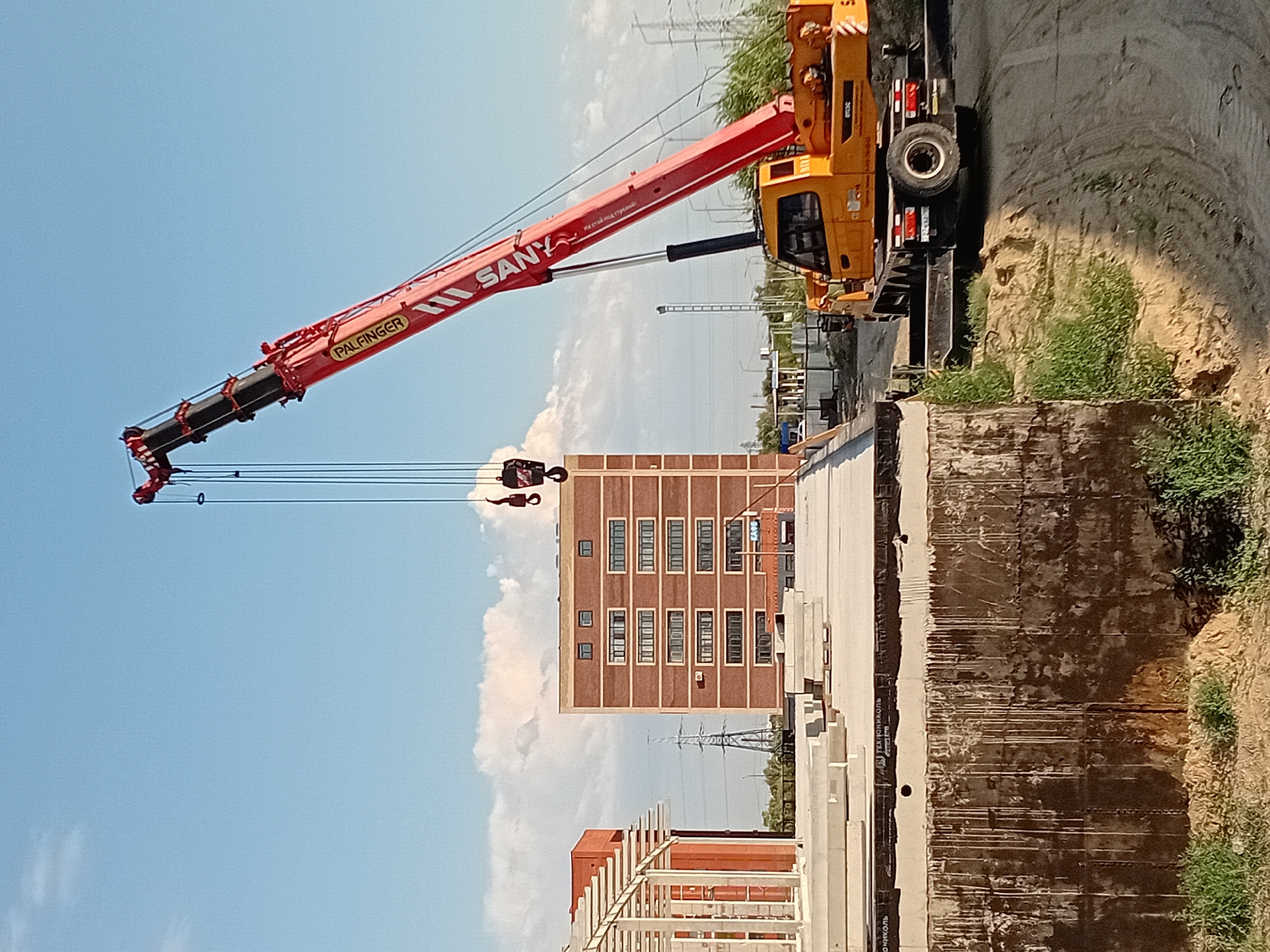 In the city of Semey, Kazakhstan, on a construction site under a clear blue sky with scattered clouds, a large red and yellow SANY crane is actively engaged in lifting operations. The crane's boom extends high into the air, with multiple cables and a hook system suspended from it. In the foreground, a concrete structure, possibly a foundation or a lower level of a building, is visible, with dark, water-stained concrete walls. To the left, a red brick building with white framing is partially obscured. In the background, a tall brick building with numerous windows stands prominently. To the right of the brick building, a yellow mobile crane is positioned on the ground, appearing to be part of the construction effort. The surrounding environment includes mounds of dirt and sand, patches of green grass, and power lines and towers in the distance, indicating an urban or semi-urban setting. The scene captures a moment of active construction work.