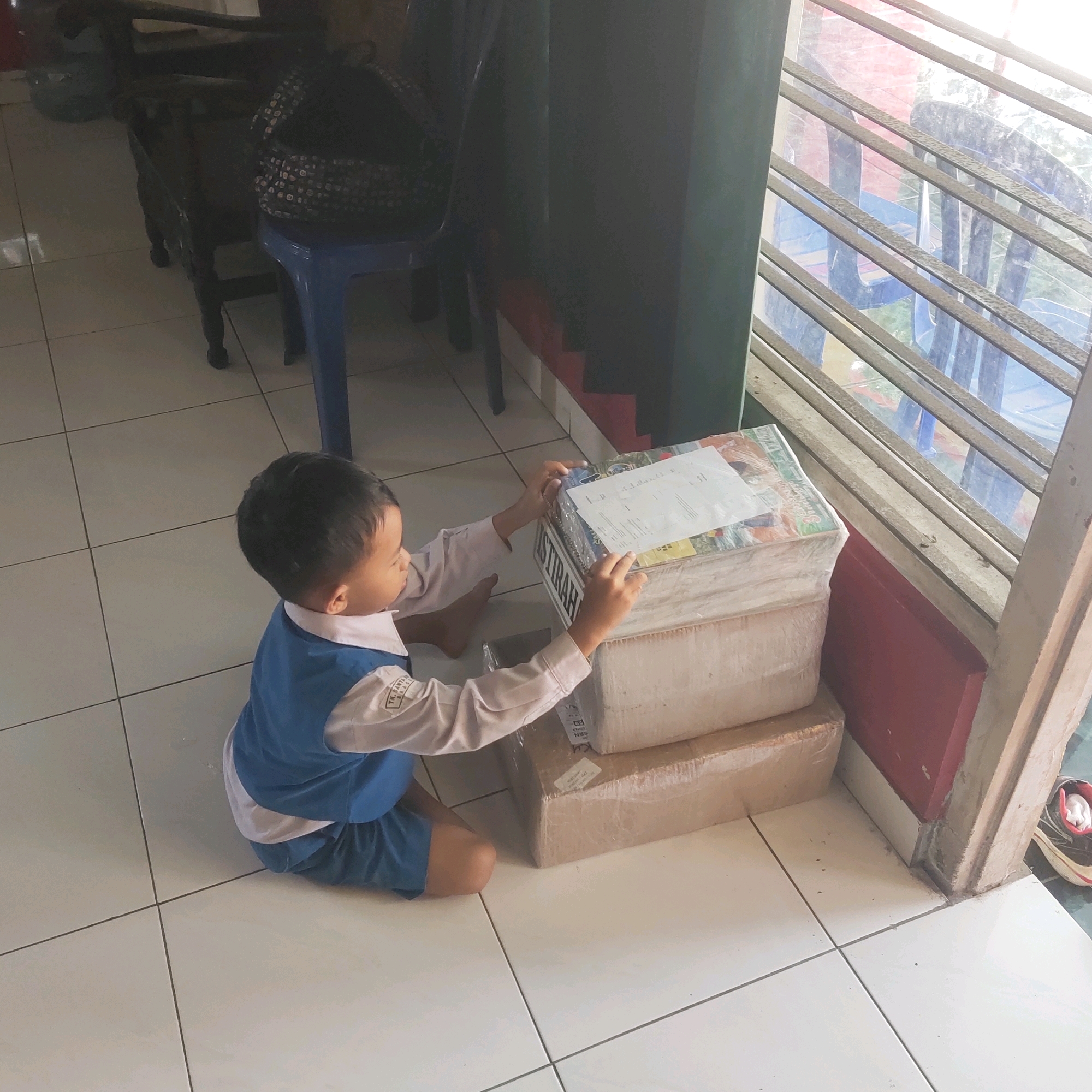 A young child wearing a blue and white uniform kneels on a tiled floor, interacting with two stacked cardboard boxes. The boxes appear to be wrapped in clear plastic and tape. The child is looking down at the boxes, with their hands resting on the top box. The child's uniform has a logo on the sleeve, indicating they attend "TK SANTA MARIA SRAGEN," a kindergarten in Sragen, Indonesia. The room has a window with metal bars, suggesting it might be a school or a residential building. There are also some chairs and other furniture visible in the background, but they are out of focus. The lighting in the image is bright, with sunlight coming in through the window.