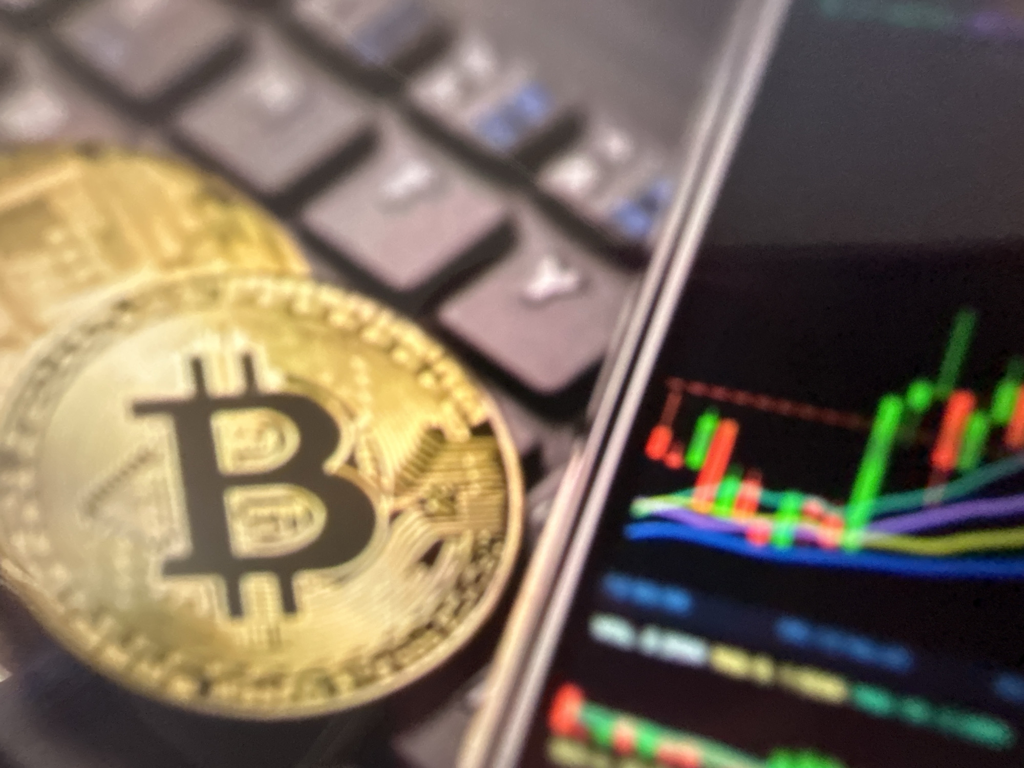 The image displays a close-up, blurred view of a Bitcoin cryptocurrency coin placed on a keyboard. To the lower left, a portion of a screen shows a cryptocurrency trading chart with colorful lines representing price movements. The scene appears to be set indoors, with artificial lighting. There is no indication of a specific location in Gelendzhik, Russia, or any people present.
