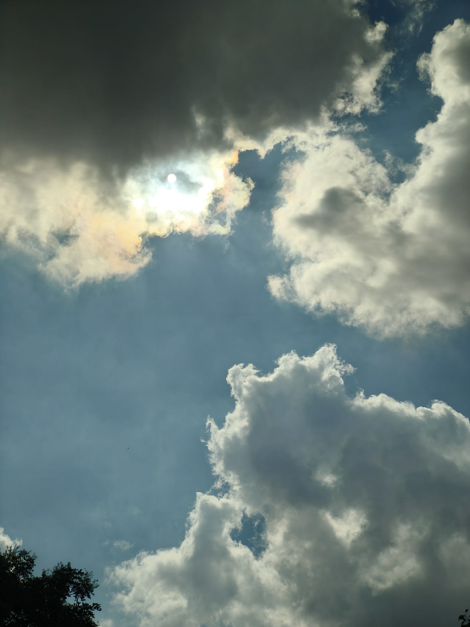 The image captures a sky with scattered clouds, and a portion of the sun is visible through a gap in the clouds, creating a bright halo effect with hints of rainbow colors. The sky is a mixture of blue and gray tones, with some clouds appearing dark and heavy, while others are bright and fluffy. In the lower left corner, the dark silhouette of tree branches is visible, suggesting an outdoor scene. The image does not depict any specific location within the city of Nanguan District, China, nor does it contain any people, subjects, objects, or actions. The scene is a natural environment focused on the atmospheric conditions.