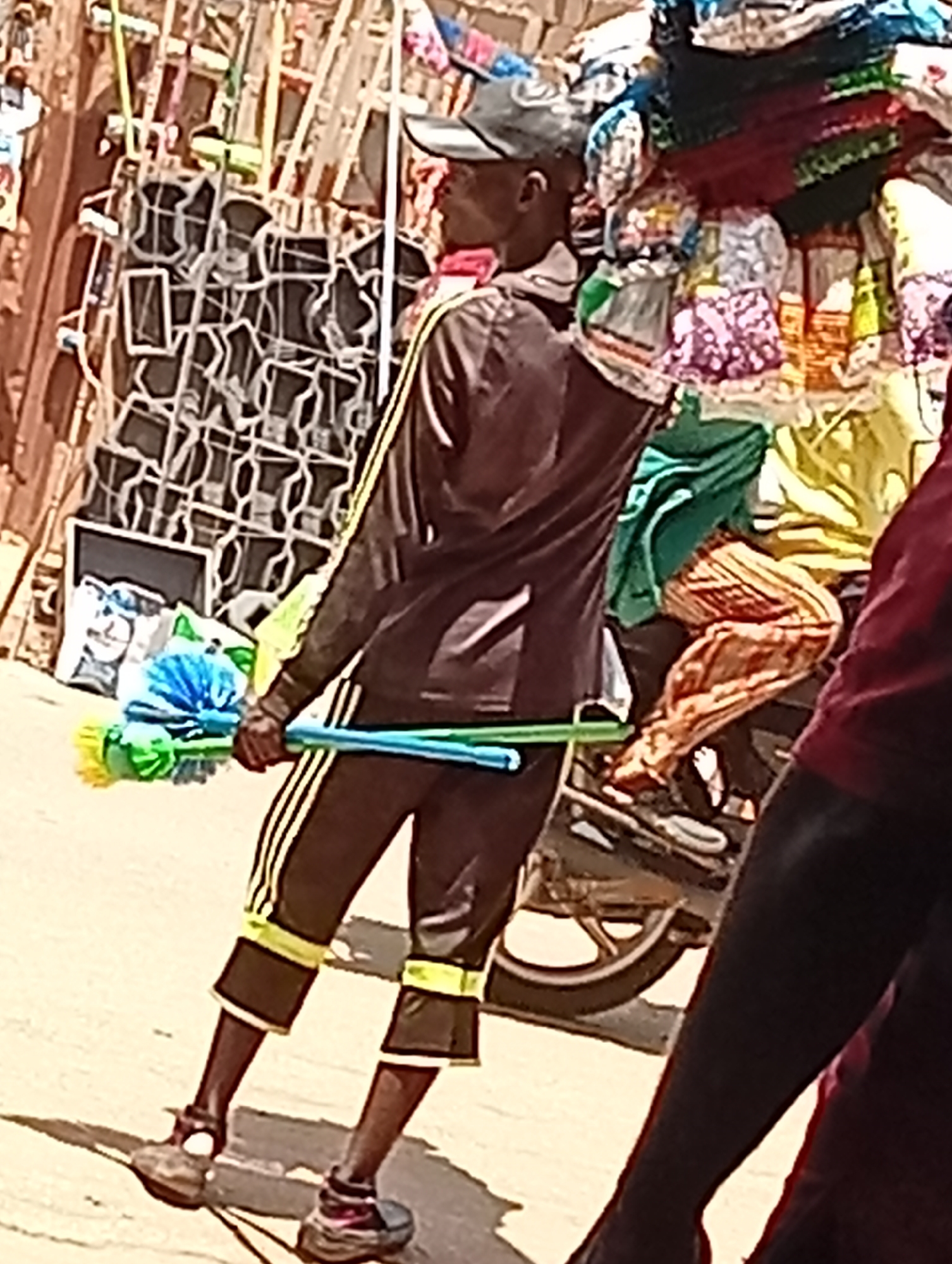 In Maiburiji, Nigeria, an individual wearing a dark-colored tracksuit with yellow stripes is holding a set of blue and green scrub brushes. He's standing near a motorcycle loaded with textiles, and there's a stack of metal frameworks nearby.