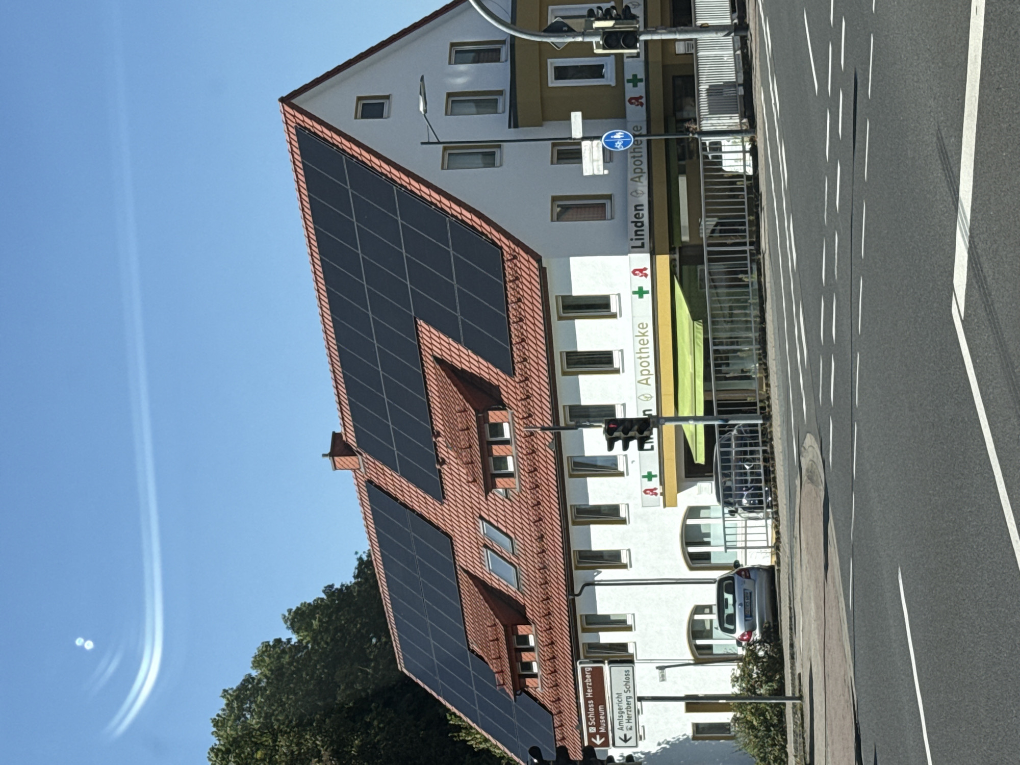 The image captures a street view in Herzberg, Germany, featuring a white building with a red-tiled roof covered in black solar panels. The building appears to be a commercial establishment, with signage indicating "Linden Apotheke" (Linden Pharmacy) and another "Apotheke" with a green awning. Two cars are visible parked in front of the building: a silver hatchback and a dark-colored vehicle. A street sign indicates directions to "Schloss Herzberg Museum" and "Amtsgericht Herzberg Schloss." Traffic lights and street lamps are present, suggesting an urban or semi-urban environment. The sky is clear and blue, with some light atmospheric streaks. The foreground shows a paved road with lane markings, indicating a crossroads.