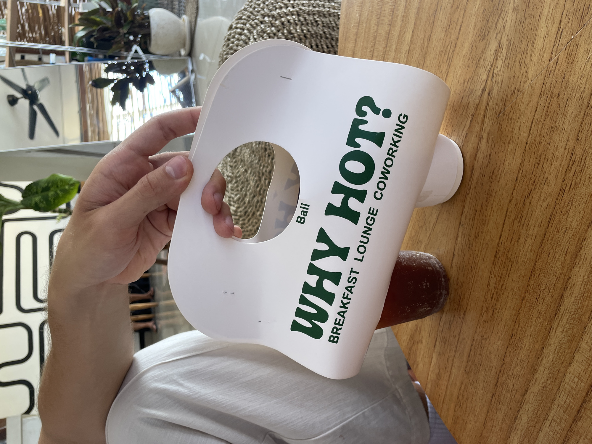 A person's arm and hand are holding a white paper drink carrier with green text that reads "WHY HOT? BALI BREAKFAST LOUNGE COWORKING." The carrier is positioned over a clear plastic cup filled with a dark liquid, likely iced coffee or tea, sitting on a wooden table. In the background, a portion of a white t-shirt is visible on the person's shoulder. Further back, a mirror reflects a ceiling fan and plants. The scene appears to be indoors, possibly in a cafe or coworking space, given the text on the carrier. The location is specified as Mengwi, Indonesia.