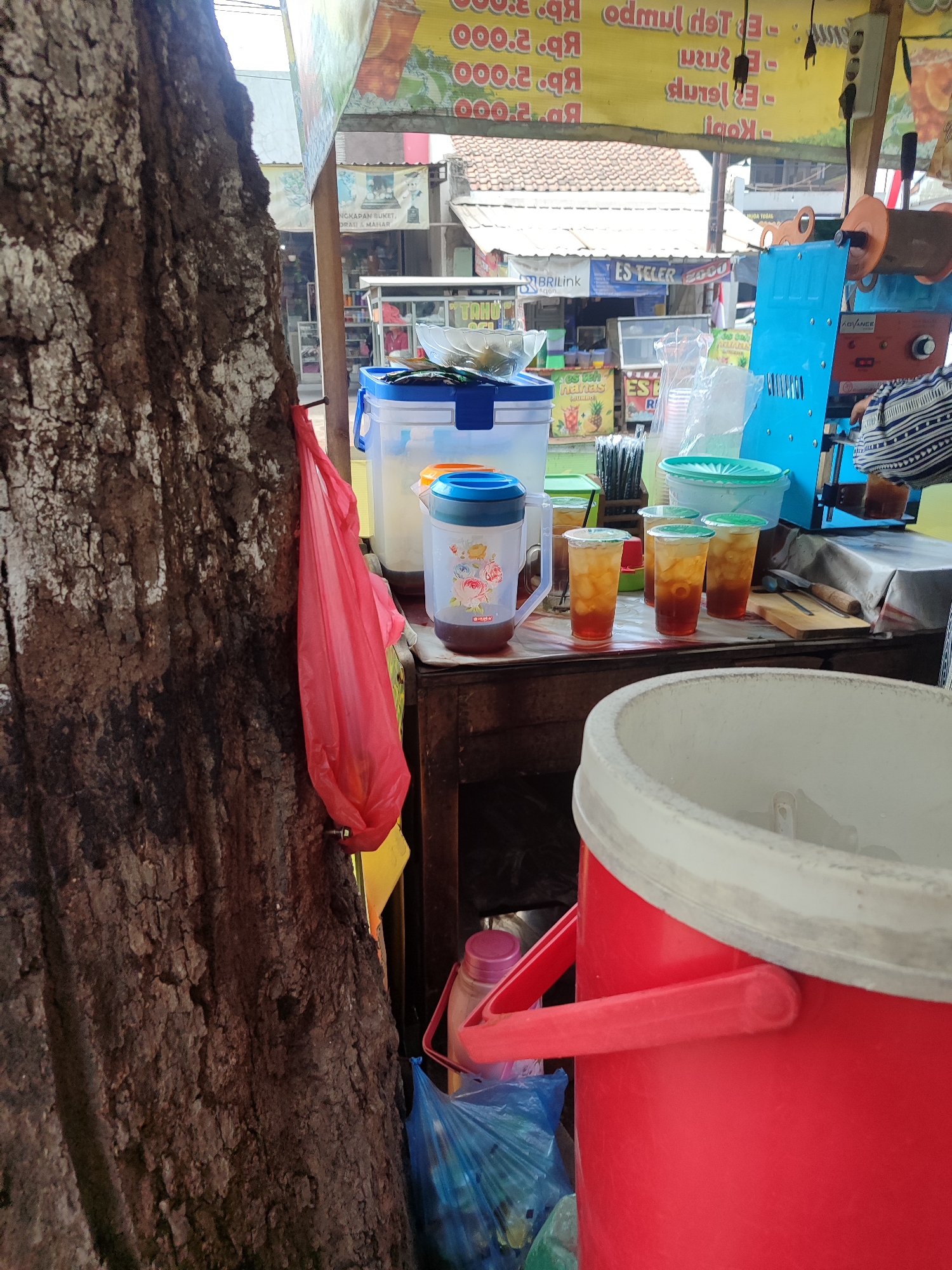 This image depicts a street food stall in Kagok, Indonesia, specializing in beverages. A large, rough-barked tree trunk dominates the left side of the frame, with a red plastic bag hanging from it. The stall itself features a variety of containers for drinks, including large plastic dispensers, some filled with iced tea. Several plastic cups filled with iced tea are arranged on a table, alongside other beverage-related items like straws and a blue ice dispenser. In the background, a woman in a striped shirt is visible, presumably serving customers. The stall appears to be set up under an awning, with signs and merchandise displayed. The overall atmosphere suggests a busy, everyday scene at a local market or roadside eatery.