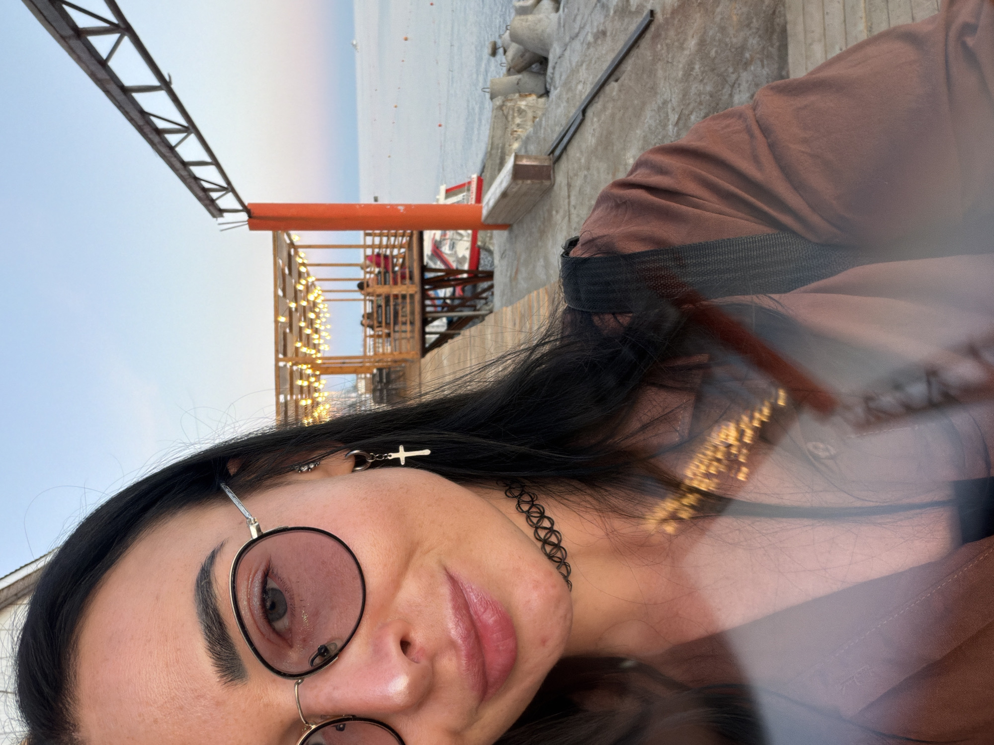 A woman is taking a selfie outdoors at dusk. She is wearing round, tinted glasses and a cross earring. A black choker is around her neck. She is wearing a brown shirt with a black strap visible over her shoulder. In the background, there is a wooden pier with lights strung across it, and a body of water with a boat. The sky is a pale blue with hints of pink.