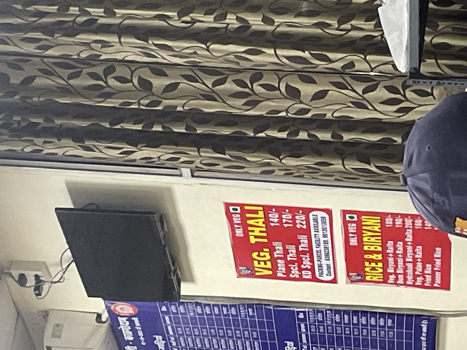 The image depicts an indoor scene, likely a casual eatery or food stall, in Haldwani, India. The upper portion of the frame is dominated by a patterned curtain featuring a repeating brown leafy design against a lighter background.
Mounted on a light-colored wall are two prominent red and white menu boards. The first board, labeled "ONLY VEG" and "VEG. THALI," lists "Plane Thali" for 140/-, "Spcl. Thali" for 170/-, and "KD Spcl. Thali" for 220/-. It also states "PACKING-PARCEL FACILITY AVAILABLE" and provides contact numbers: 6388238189 and 9012613858. The second board, also marked "ONLY VEG" and titled "RICE & BIRYANI," offers "Veg. Biryani+Raita" for 180/-, "Dum Biryani+Raita" for 190/-, "Hydrabadi Biryani+Raita" for 200/-, "Veg. Pulao+Raita" for 160/-, "Fried Rice" for 140/-, and "Paneer Fried Rice" (price partially obscured).
To the left of the menu boards, a black rectangular device, possibly a small monitor or speaker, is mounted on the wall, with visible electrical wiring and an outlet below it. Beneath the red menu boards, a larger blue and white information or menu board is partially visible, with the text "श्री - काठगोदाम" (Shree - Kathgodam) at its top left corner.
On the far right, part of a person's head, wearing a dark blue baseball cap, is visible, suggesting a customer or staff member is present in the scene. The overall ambiance indicates a functional establishment offering vegetarian meals.