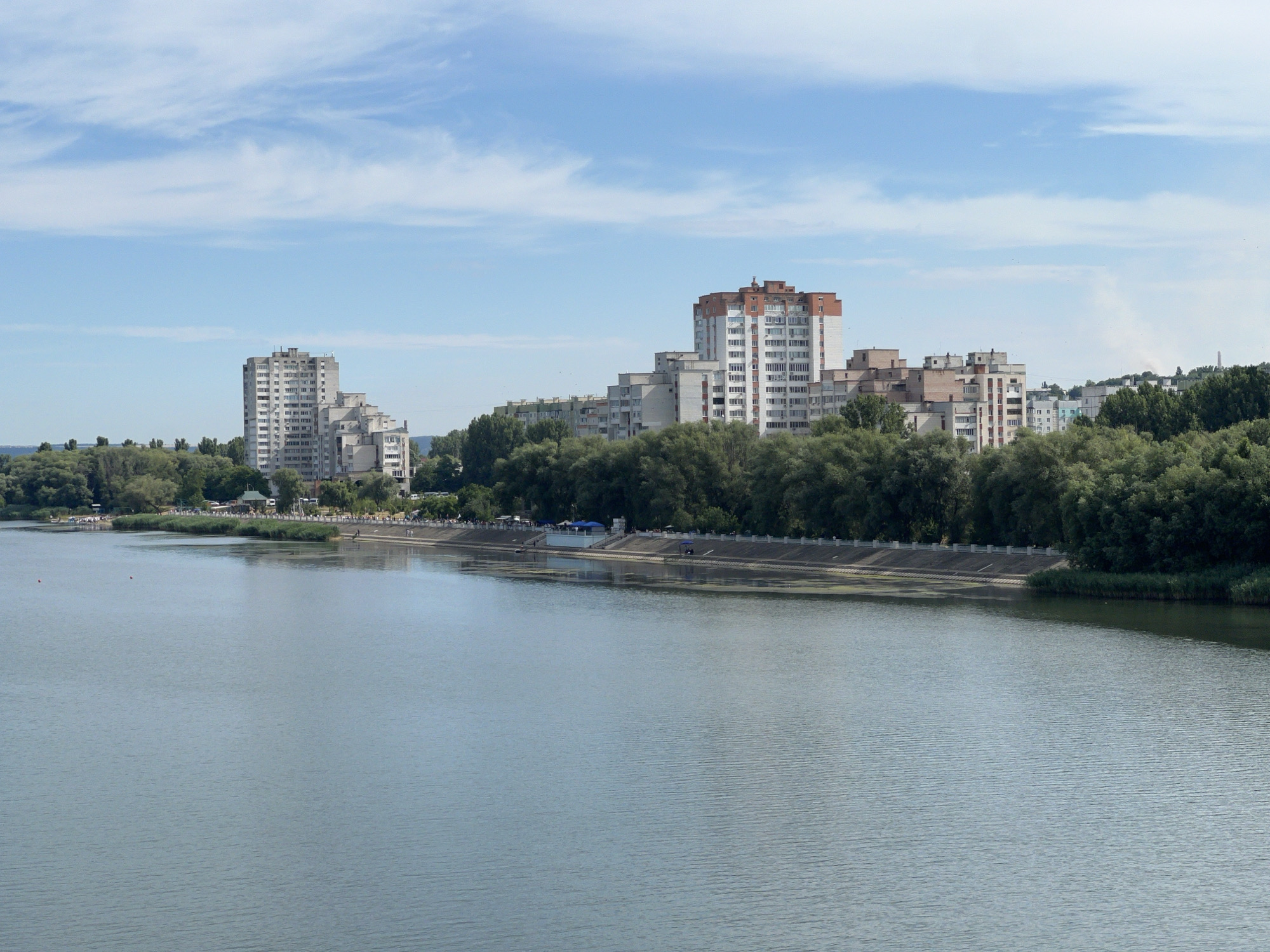 The image captures a wide, calm body of water, likely a river, with a city skyline visible on the opposite bank. The foreground is dominated by the textured surface of the water, reflecting the soft light of a partly cloudy sky. Along the far bank, a dense line of green trees forms a natural barrier separating the water from the urban landscape. Behind the trees, a series of multi-story buildings rise, characterized by varying architectural styles and colors, with a prominent red-and-white building standing out. A paved walkway or embankment runs along the water's edge, with scattered individuals and small groups of people visible, some standing and others appearing to be walking. There are also a few blue umbrellas suggesting a recreational area or temporary stalls. The overall scene depicts a waterfront area within a city, likely Rezina, Moldova, showcasing a blend of natural greenery and residential development. The sky is a pale blue with streaks of white clouds, indicating fair weather.