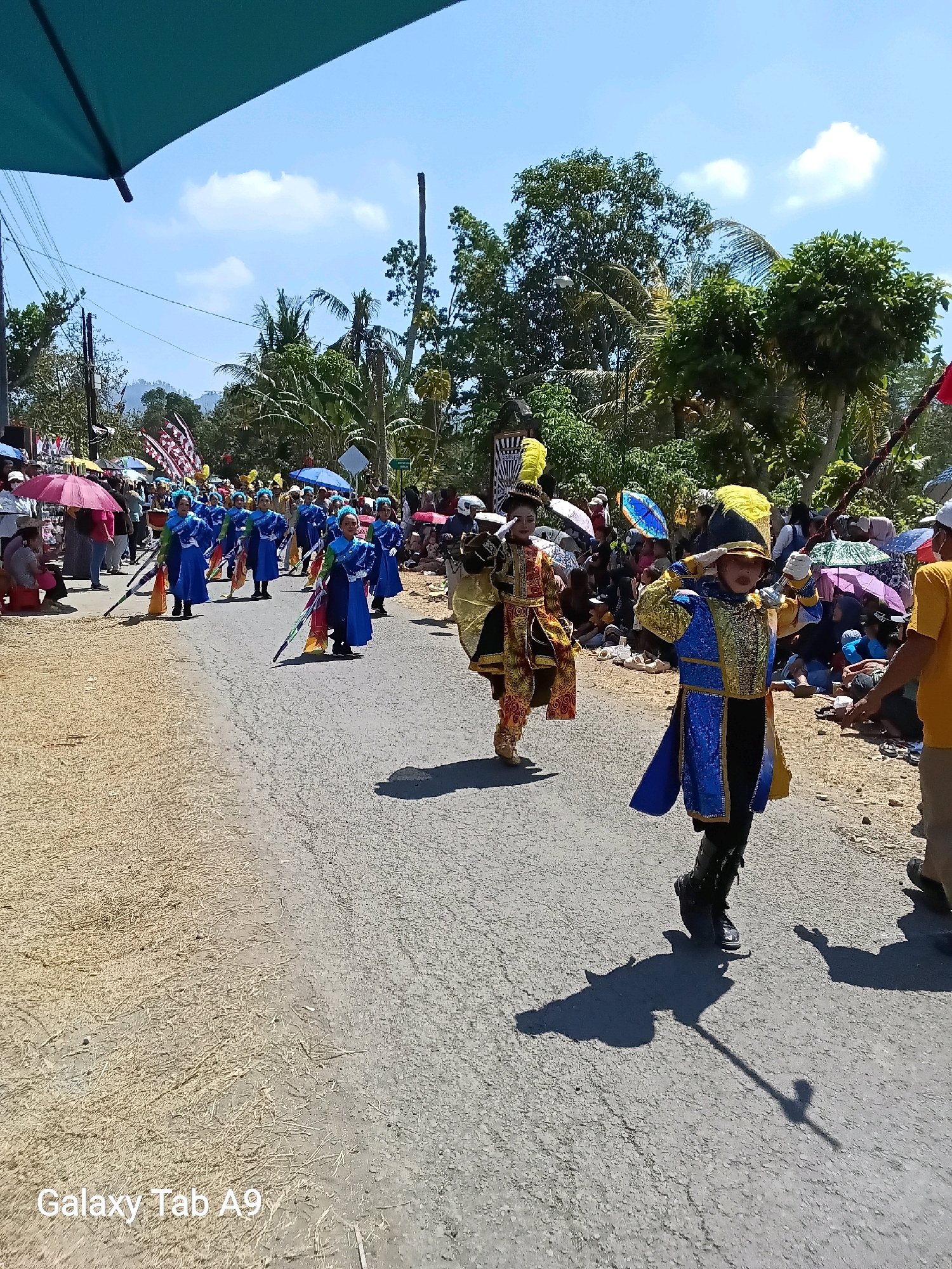 This is a medium shot of a parade in Ngadirejo, Indonesia. In the foreground, a person in a blue and gold marching band uniform salutes the viewer. They are wearing a gold helmet with a blue plume, a blue jacket with gold trim, and black pants tucked into black boots. Their shadow is cast on the asphalt road. Behind them, another person in a similar, but different colored uniform, is also marching. In the background, a group of children in blue marching band uniforms are walking in formation, holding flags. Spectators are lined up on the side of the road, some holding umbrellas for shade. The parade is taking place on a sunny day, with a blue sky and scattered clouds. Trees and buildings can be seen in the background, suggesting a rural or semi-urban setting. The ground is a mix of asphalt and dry grass or straw.