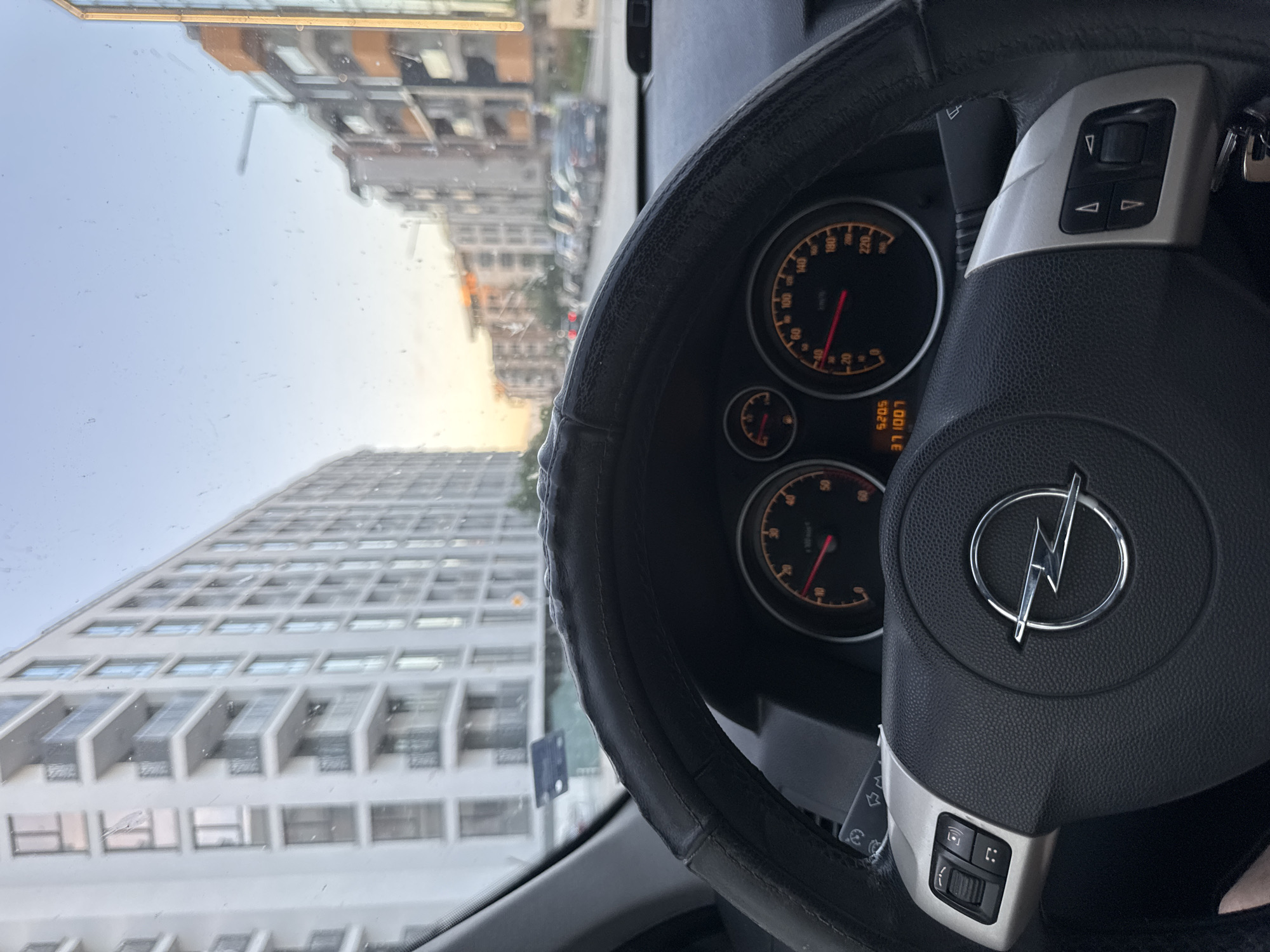 The image, taken from the interior of an Opel car, shows the vehicle’s steering wheel and dashboard in the foreground. The speedometer needle reads approximately 15 km/h, and the odometer displays “371007 km”. The Opel logo is prominently displayed on the steering wheel.
Looking through the windshield, a cityscape is visible, including a tall, white high-rise building with repeated window units, suggesting an apartment or office structure. Other buildings of varying heights and styles line the street in the distance. The sky is pale, indicating dawn or dusk.
The location is Sofia, Bulgaria.