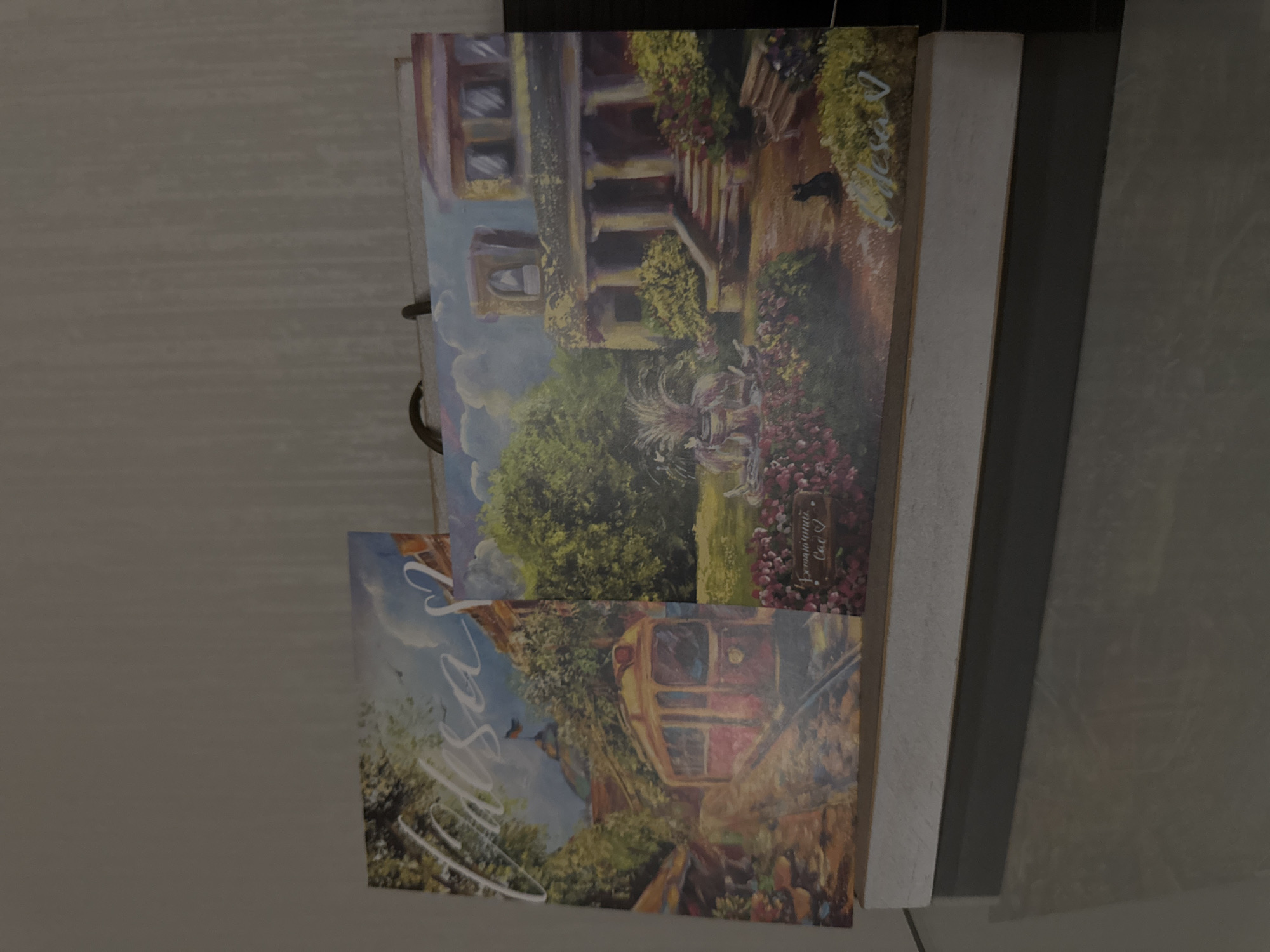 An image shows two paintings resting on a surface. The painting on the left depicts a red tram on a cobblestone street with buildings and trees in the background. The text "Odesa" is written in white script in the upper left corner. The painting on the right features a large house with columns and a garden with flowers, a fountain, and a black cat. Text in white script is visible at the bottom of this painting. Both paintings appear to be displayed together, possibly as part of a set.