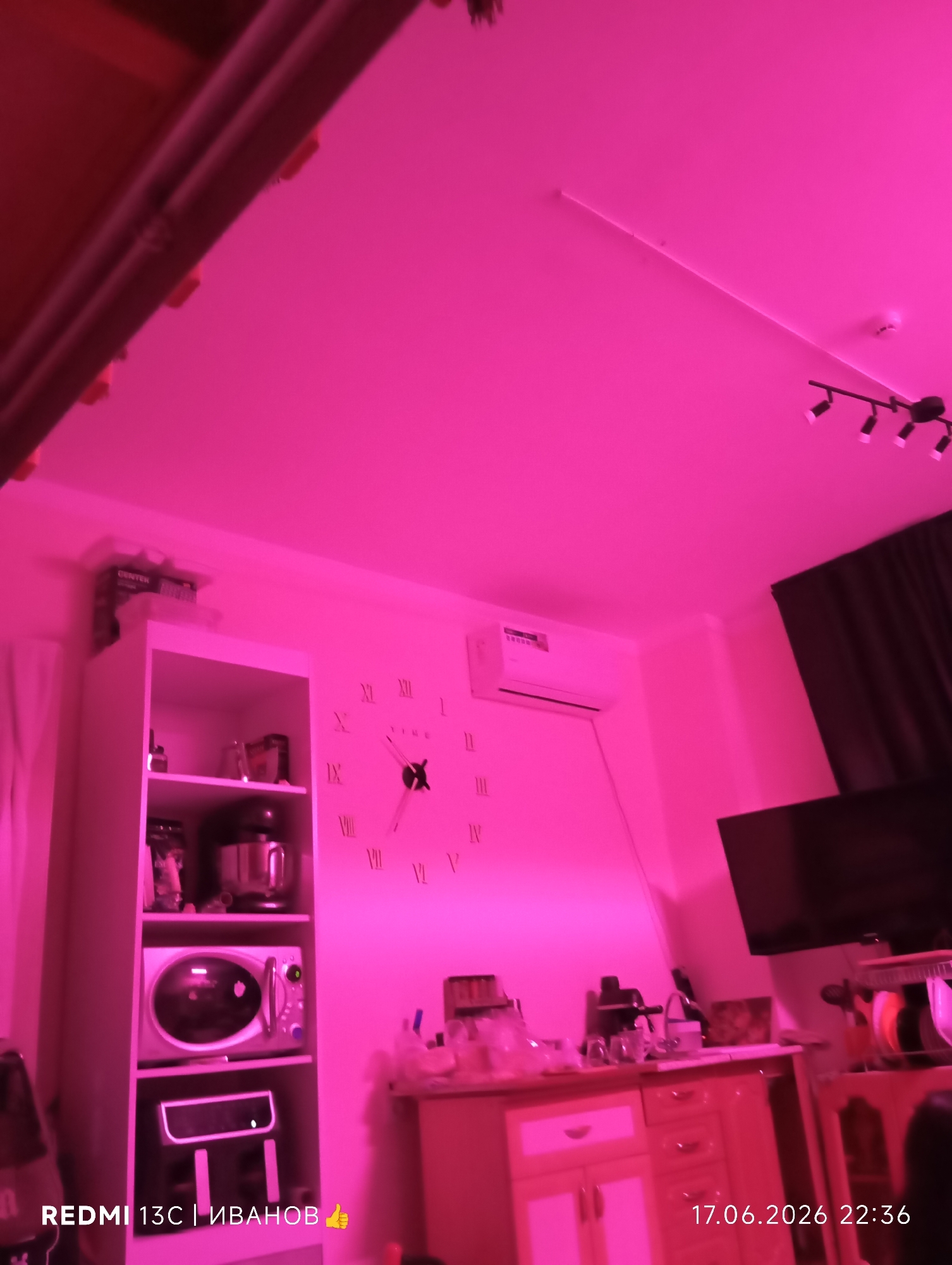 The image depicts an indoor scene illuminated by pink lighting, likely from LED strip lights. The room appears to be a kitchen or dining area. In the foreground, a tall white cabinet is visible, filled with various appliances and kitchenware. These include a stand mixer, a microwave, and what seems to be an air fryer or similar appliance. On a shelf above, there are boxes, one with the label "GORENJE".
To the right of the cabinet, a large clock with Roman numerals is mounted on the wall. The clock hands are visible. Further to the right, a black curtain hangs, and above it, a dark flat-screen television is mounted. Below the television, a light-colored cabinet with drawers and doors is present. On top of this cabinet, several items are arranged, including what looks like a coffee maker and glasses.
In the upper part of the image, an air conditioning unit is mounted on the wall, and a set of three spotlights is attached to the ceiling.
Visible text includes "REDMI 13C | ИВАНОВ 👍" in the bottom left corner, indicating the phone model and likely the name of the person who took the photo. In the bottom right corner, the date and time "17.06.2026 22:36" are displayed. The overall impression is that of a domestic space, possibly at night, given the late hour. The pink lighting creates a somewhat unusual and atmospheric ambiance. There are no people visible in the image. The location context of Nyurba, Russia, does not offer specific visual cues within the image itself.