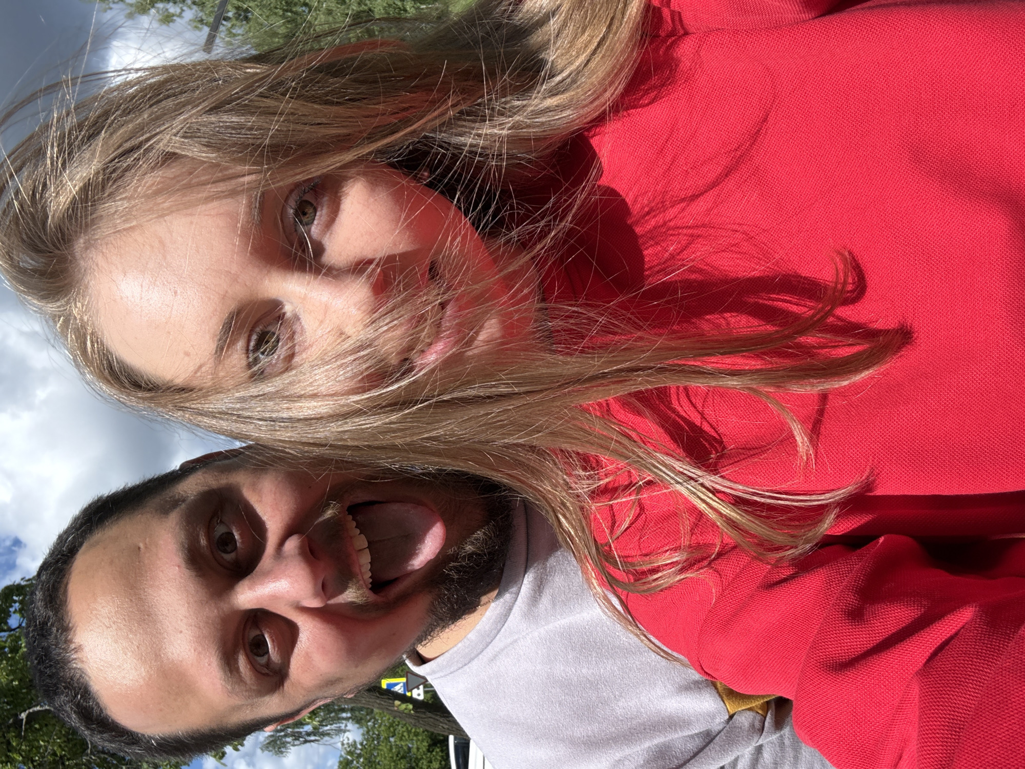 Two individuals are captured in a selfie-style photograph. A woman with blonde hair and fair skin is wearing a red garment. A man with dark hair and a beard is wearing a grey shirt and sticking out his tongue. The background shows sky, clouds and foliage, indicating an outdoor setting. The location appears to be in an unidentified urban area.