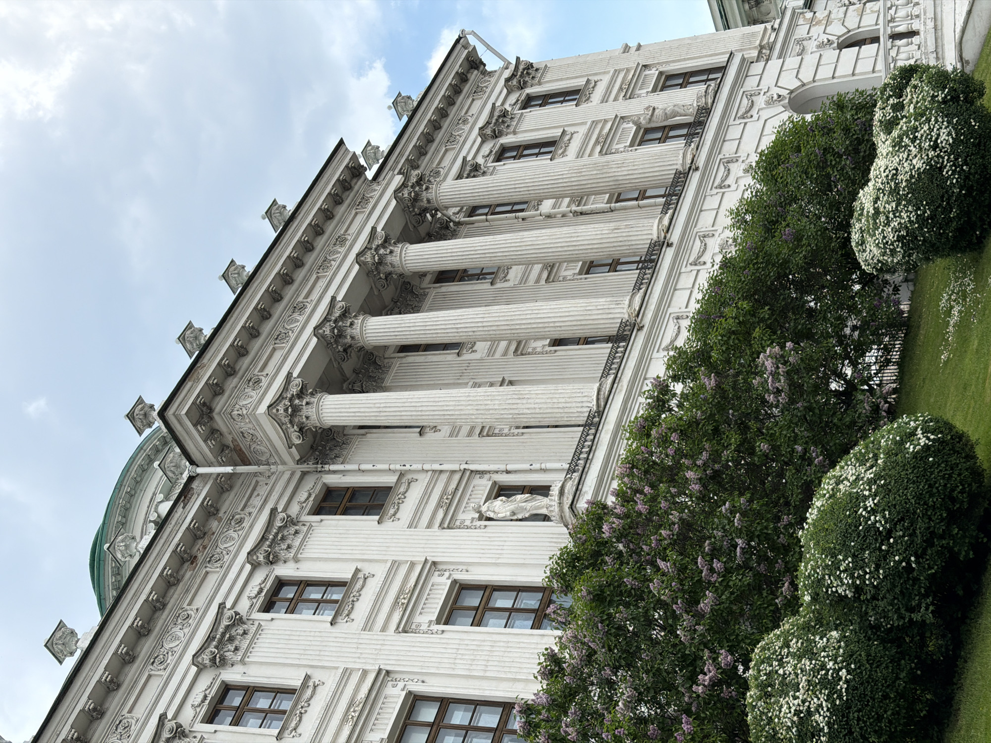 A low-angle photograph depicts an ornate, white building with multiple columns in Moscow, Russia. The architecture is heavily detailed with symmetrical patterns, statues, and moldings. Dark-framed windows are evenly spaced across the facade. In the foreground, green bushes and a lilac tree with purple flowers stand in a grassy area. The sky is partly cloudy, and the overall lighting suggests a daylight setting. A fragment of a green dome is visible on the far left.