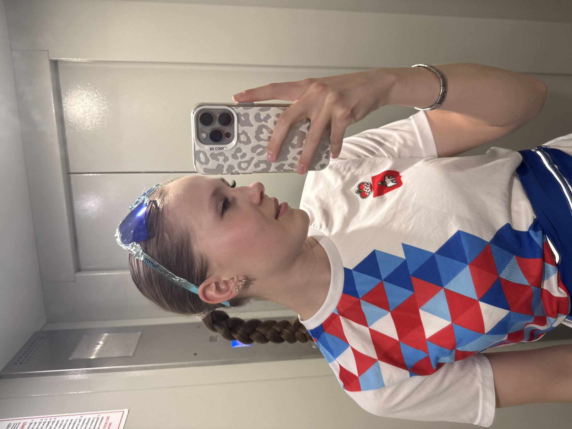 A fair-skinned young woman takes a selfie in an elevator. She is wearing a white athletic shirt with a triangular red, white, and blue geometric pattern on the left sleeve and shoulder area. There is an emblem with a crown and what appears to be a falcon or eagle on the upper right chest area of the shirt. She has braided light brown hair and blue sunglasses perched on her head. She holds an iPhone with a leopard print case. She is wearing a metal bracelet on her left wrist and multiple piercings in her left ear. The elevator has paneled walls and appears to have a metallic panel. The image may have been taken at an athletic facility in Chaikovsky, Russia, based on the shirt design.