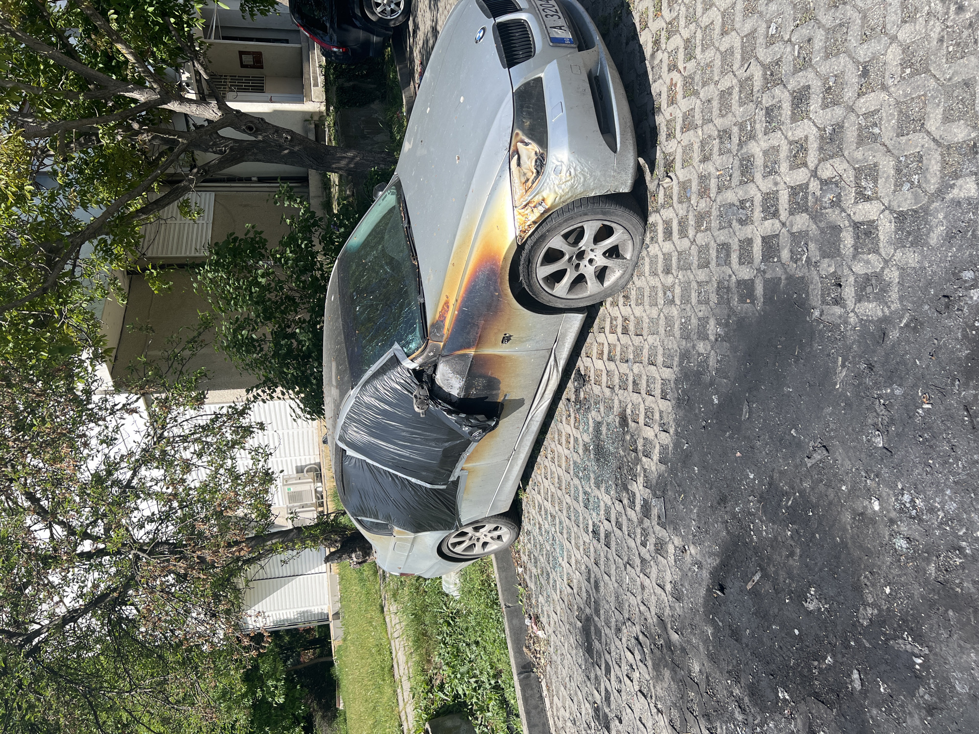 A severely fire-damaged silver BMW 3 Series sedan is parked on a paved surface in an urban area. The car's front driver's side is charred, with melted components visible. The driver's side windows are covered with black plastic. A portion of the hood and the front passenger side show signs of fire damage. The license plate, partially visible, appears to read "A 3204 BG," indicating registration in Bulgaria. Black residue is visible on the pavement near the car, suggesting the fire's origin point. Buildings and trees surround the scene. Based on the license plate, the location is likely in Burgas, Bulgaria.
