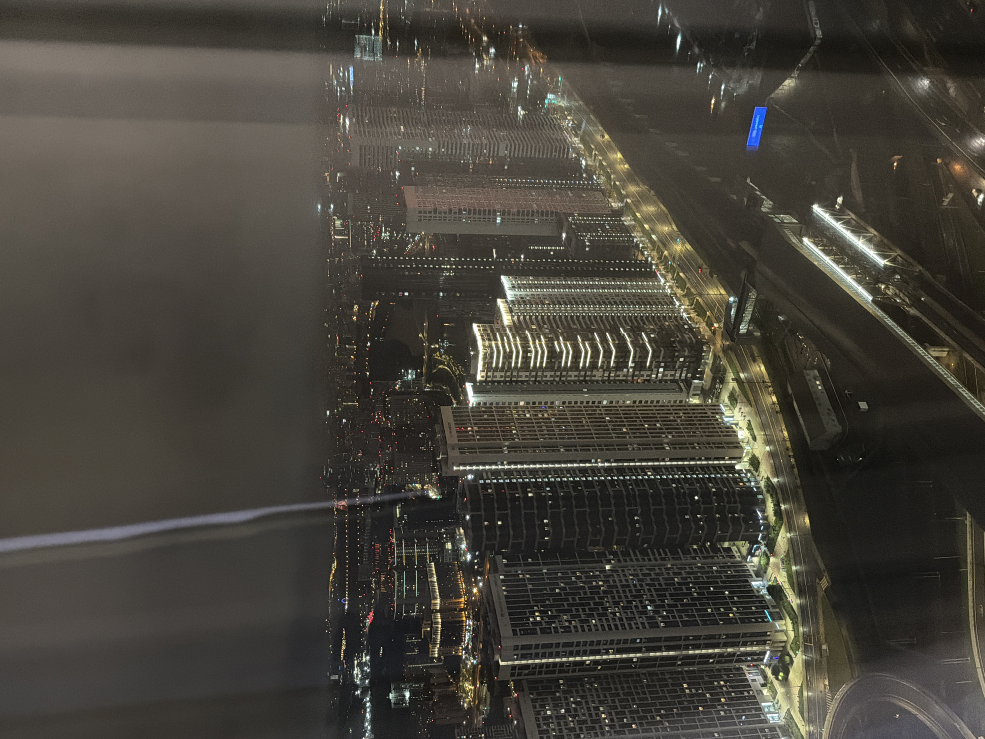 The image is an aerial, nighttime view of a modern city district, likely in Khimki, Russia, characterized by numerous high-rise apartment buildings illuminated with interior and exterior lights. A prominent vertical beam of light, possibly from a searchlight or lighthouse, cuts through the dark, cloudy sky from the upper left to the middle of the frame. In the foreground, several tall, rectangular residential buildings with patterned facades dominate the scene. A body of water, possibly a river or canal, is visible in the middle ground, reflecting the city lights. Along the waterfront, a network of roads and elevated train tracks are illuminated, with some structures appearing to be train stations or platforms. A blue illuminated sign is visible on the right side of the frame, above the train tracks. The overall impression is of a bustling, modern urban environment under a cloudy night sky.