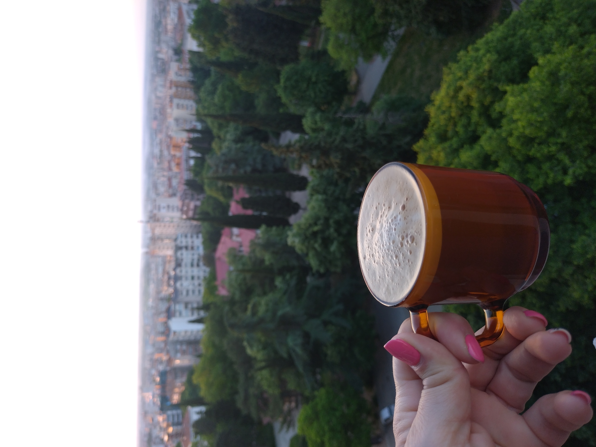 A hand holds a brown glass mug filled with a beverage, likely coffee or beer, against a backdrop of the city of Stara Zagora, Bulgaria. The hand, with pink nail polish, firmly grips the mug by its handle. The beverage has a foamy top. The city stretches out in the background, with buildings visible amongst green trees, likely viewed from a high vantage point, such as a balcony. The sky is a pale shade, suggesting either early morning or late afternoon. The overall lighting is soft and natural.