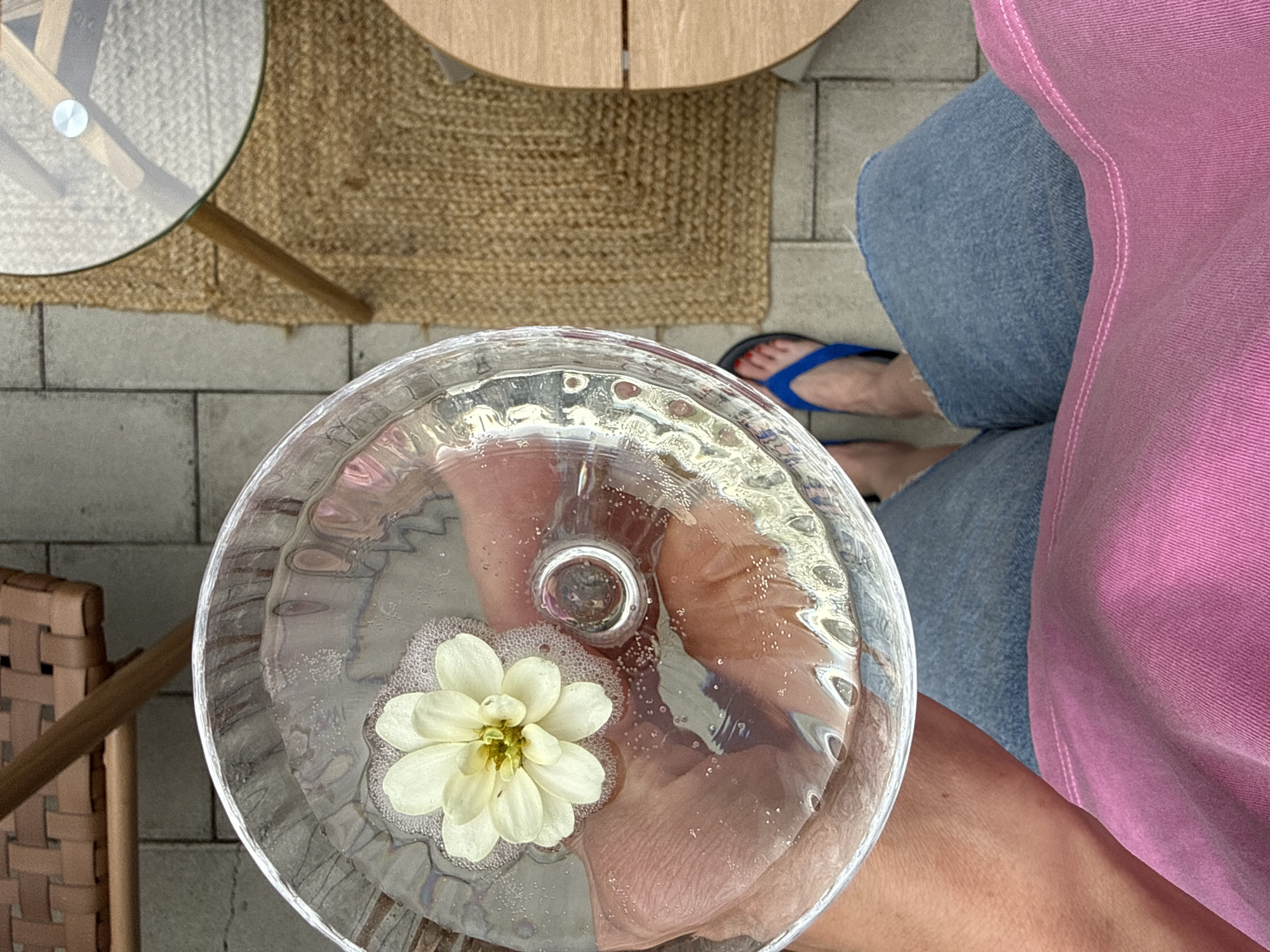 A top-down, medium shot shows a person's legs and a hand holding a glass of water with a flower in it. The person is wearing blue denim jeans and a bright pink shirt. They are also wearing blue flip-flops. The glass is clear and filled with water, and a small white flower with yellow in the center floats on top. The background appears to be an outdoor patio with stone tiles. In the background, a tan rug and a glass-topped table with wooden legs are visible. The scene suggests a relaxed outdoor setting, potentially in a cafe or residential area. The specific location of Tbilisi, Georgia is not visually evident from the image.